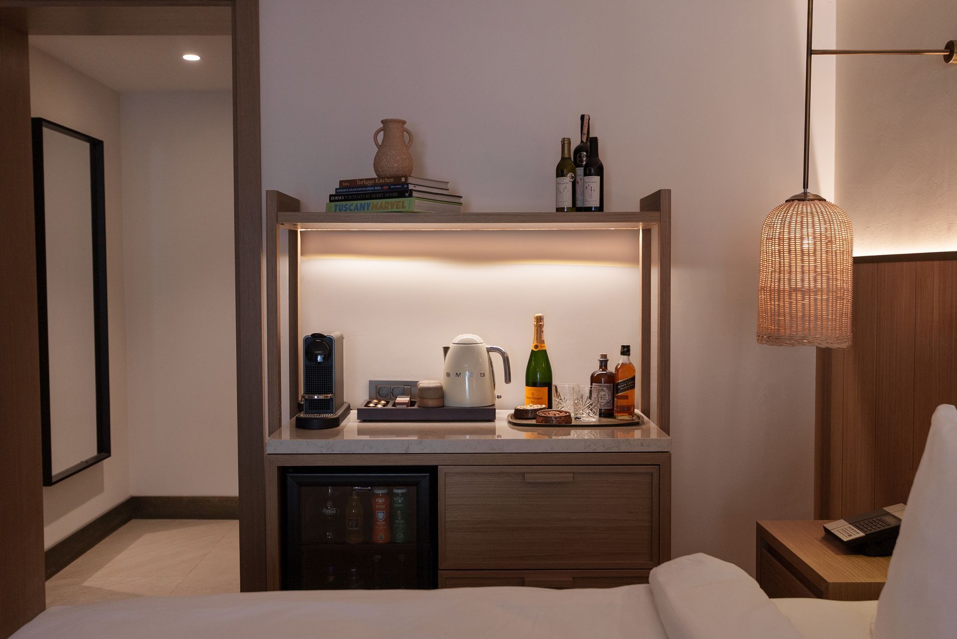 A hotel room coffee and beverage station with a small refrigerator, kettle, coffee maker, and bottles on a wooden counter.