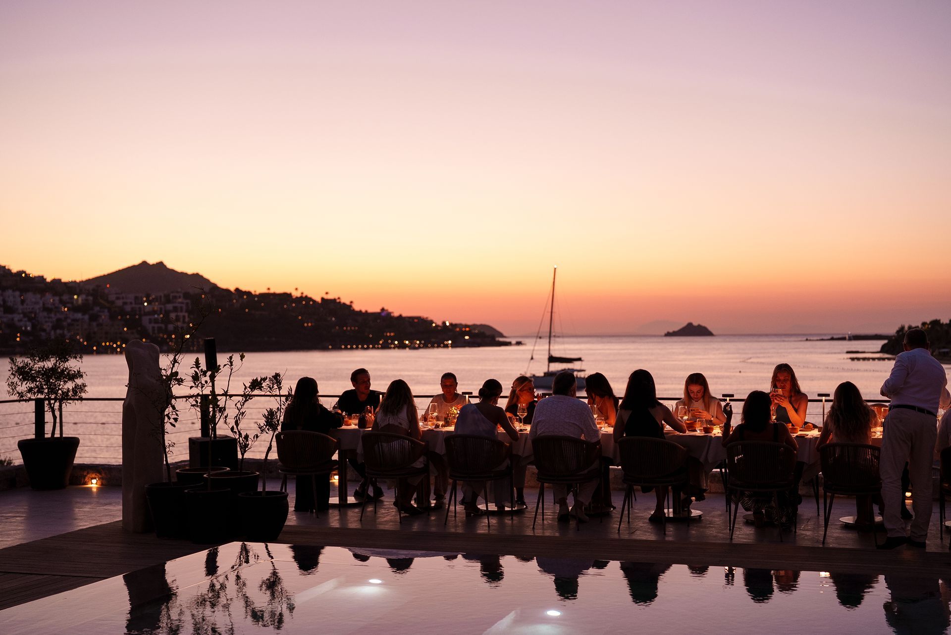 A group of people dining at a long table by the water at sunset, with their reflection visible in a foreground pool.