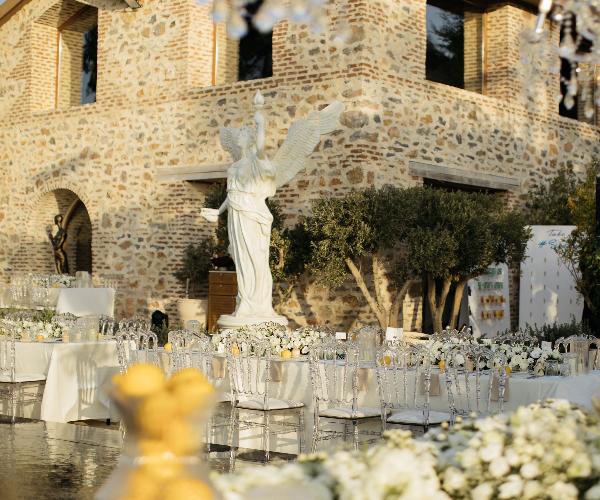 Outdoor wedding reception with white tables, transparent chairs, and a large winged statue in front of a stone building.
