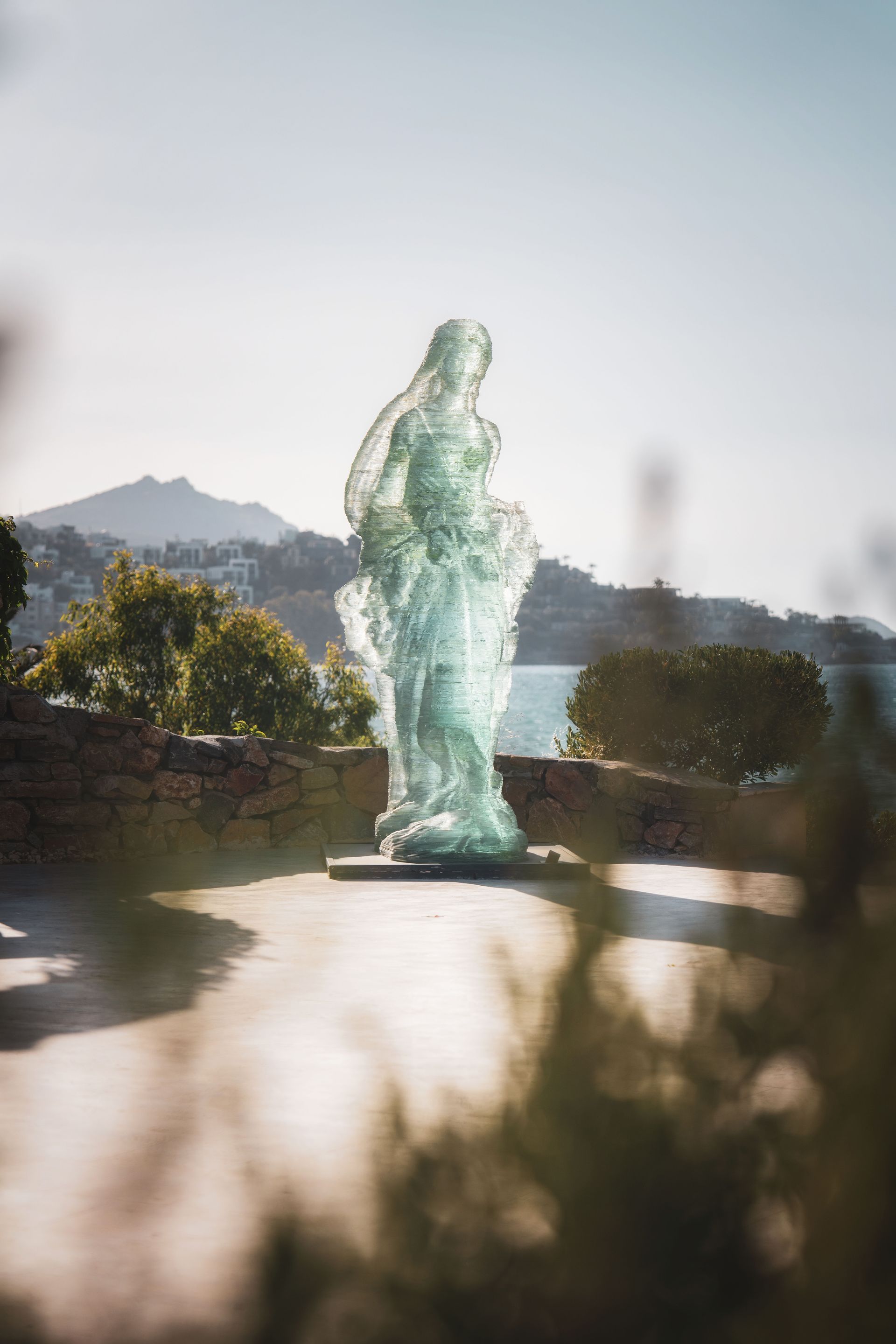 A translucent, light-green glass sculpture of a figure stands on a pedestal overlooking a sunny coastal landscape.