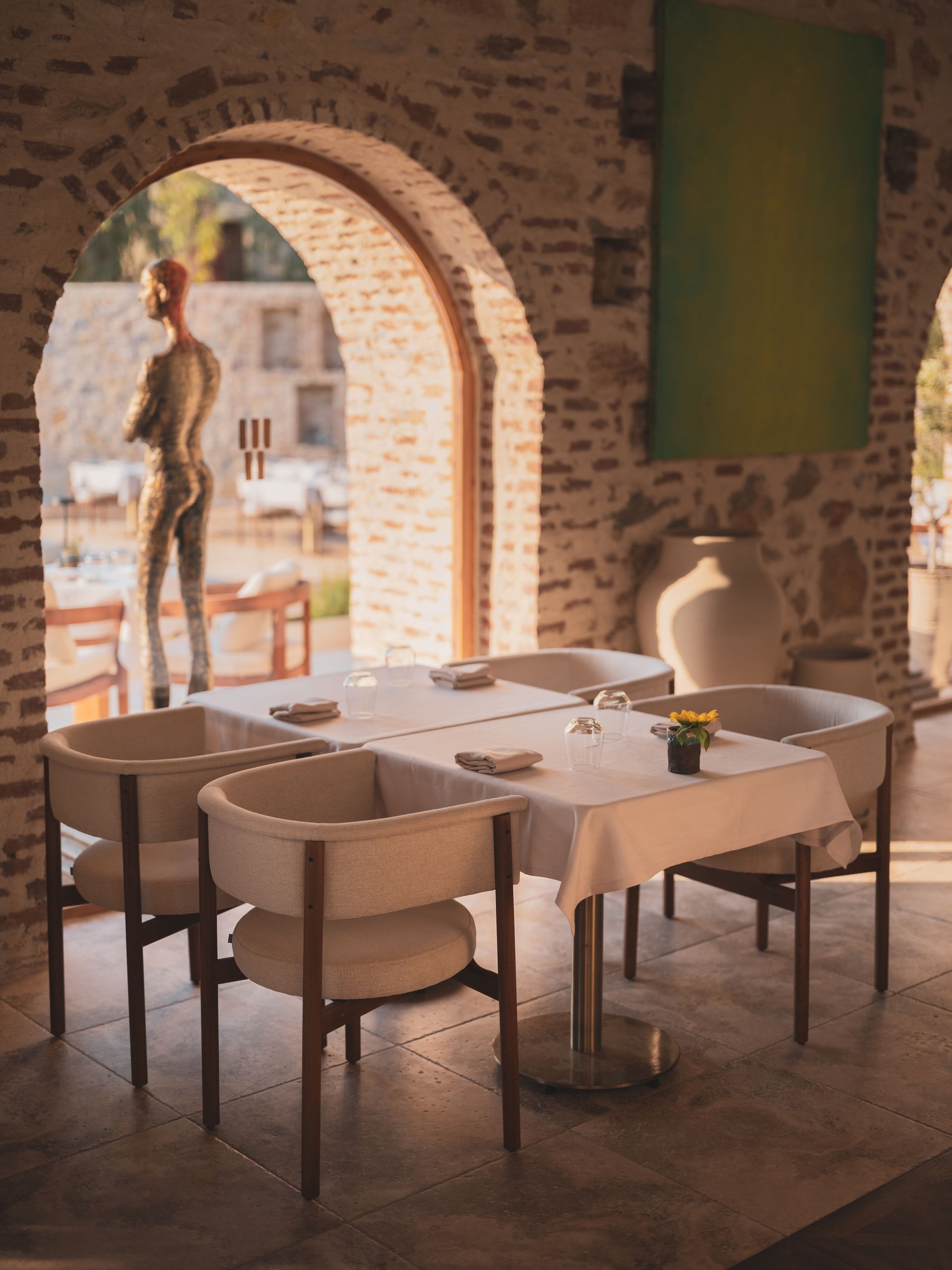 A table with four chairs set for dining inside a stone archway, with a sculpture visible in the background.