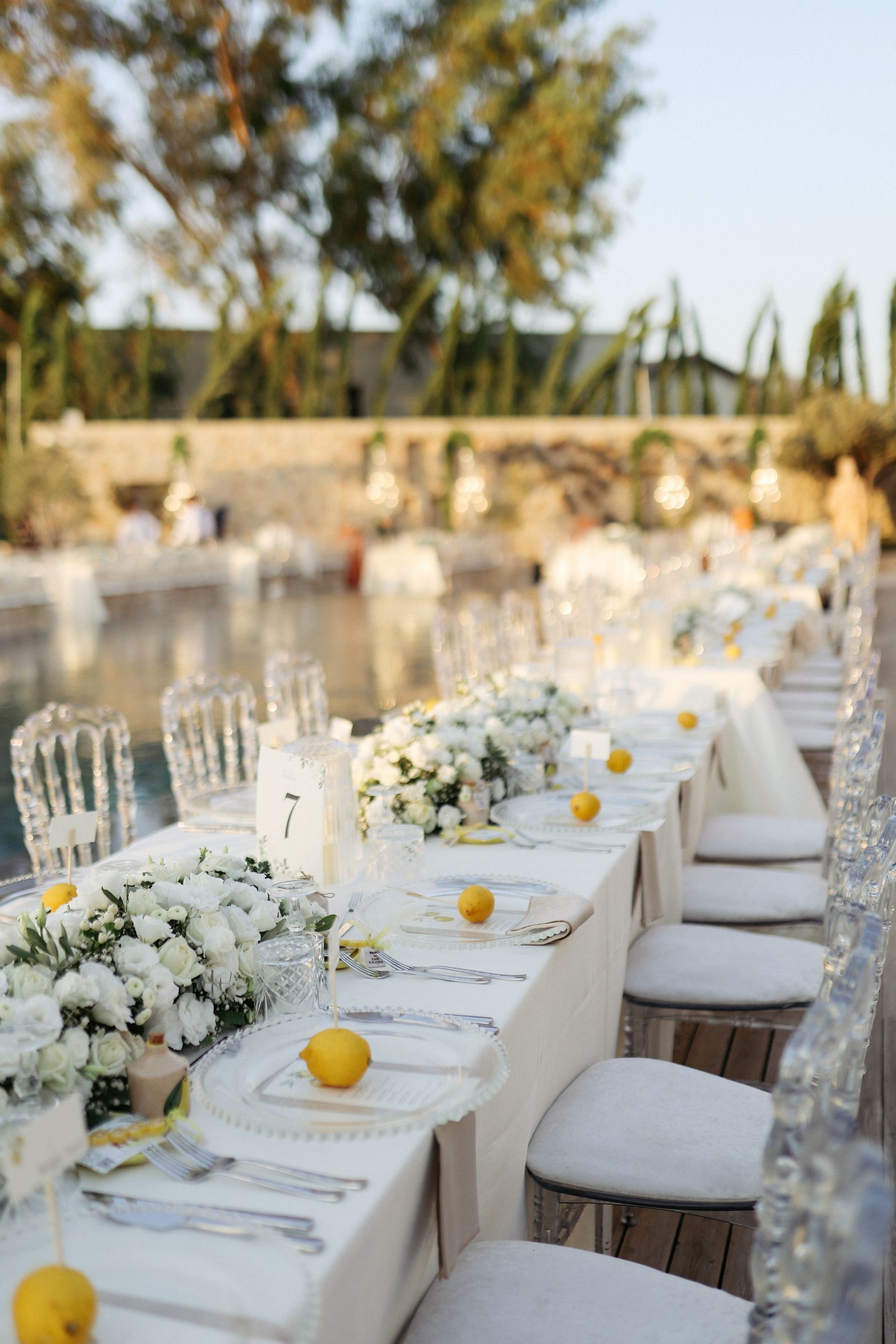 Long table set for an outdoor event with white floral arrangements, yellow lemons, and transparent chairs.