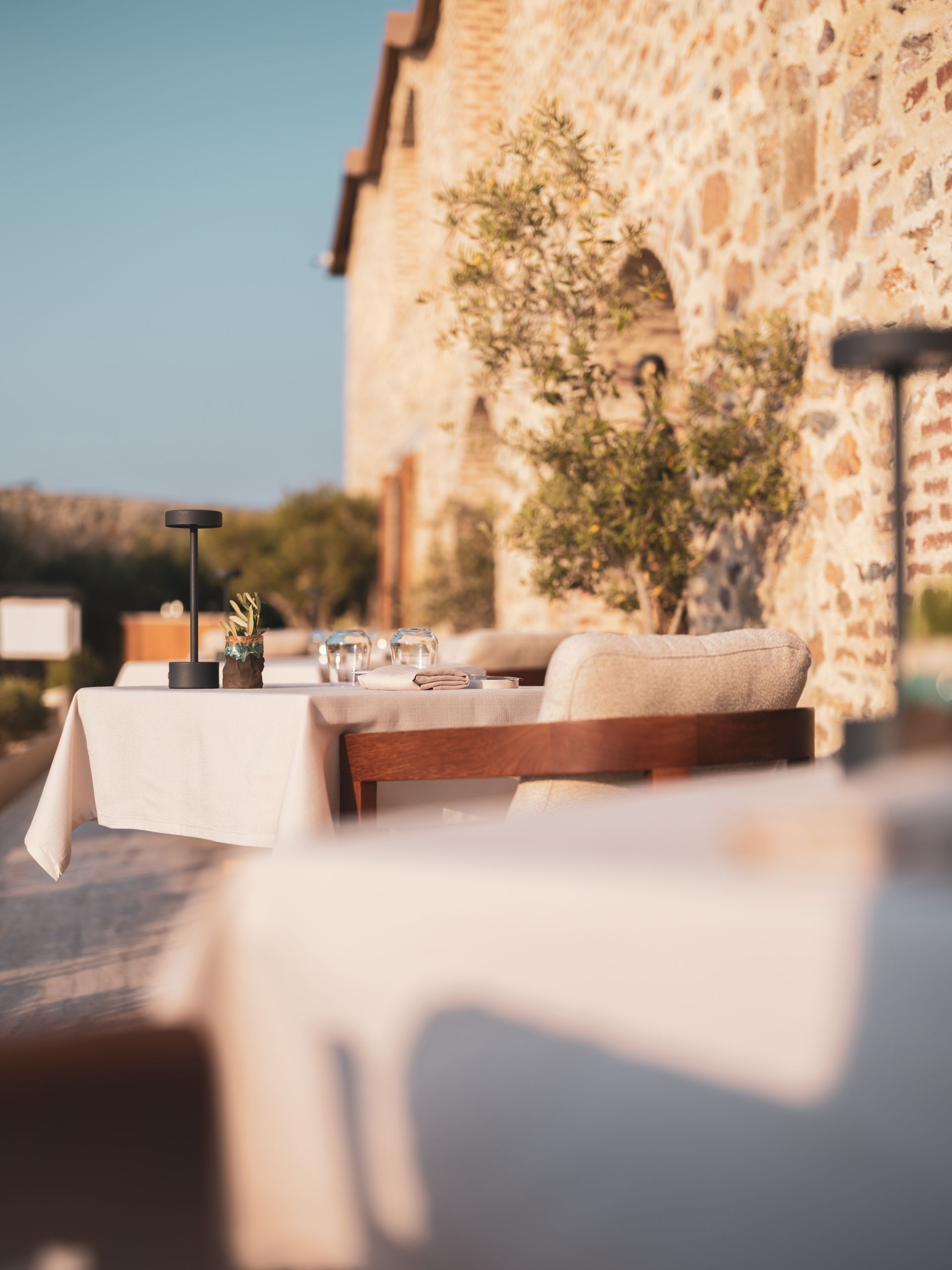 An outdoor patio dining area with white-clothed tables, chairs, and lamps against a rustic stone building at sunset.