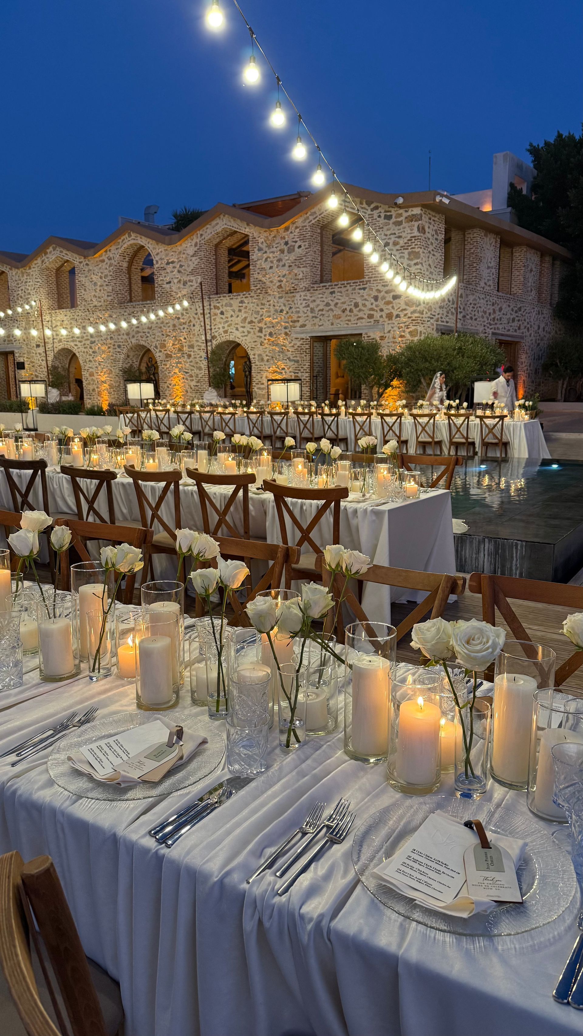 Long outdoor tables set for a wedding dinner in front of a stone building, illuminated by glowing string lights at dusk.