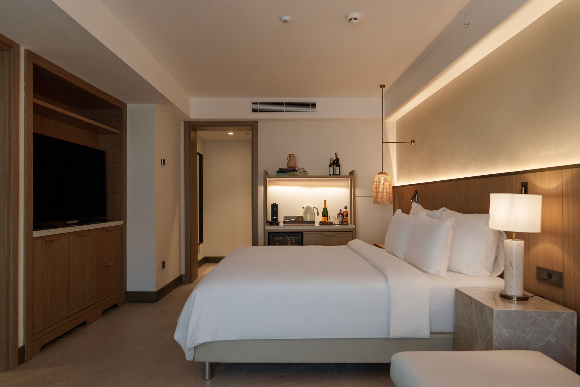 A clean, modern hotel bedroom with a large bed, wooden accents, a wall-mounted TV, and a mini-bar area.