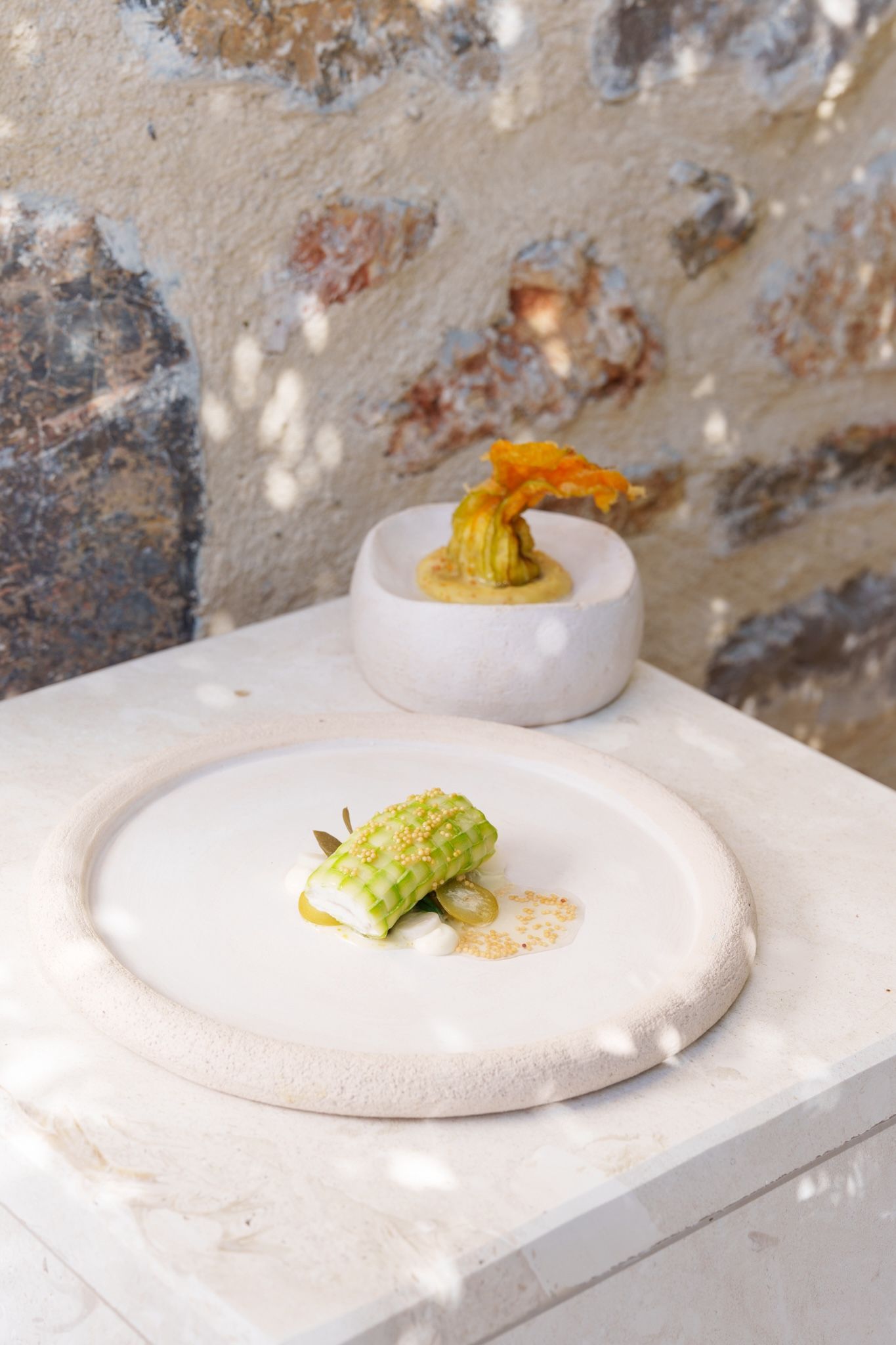 Two gourmet dishes served on textured, cream-colored ceramic plates against a rustic stone wall with dappled sunlight.