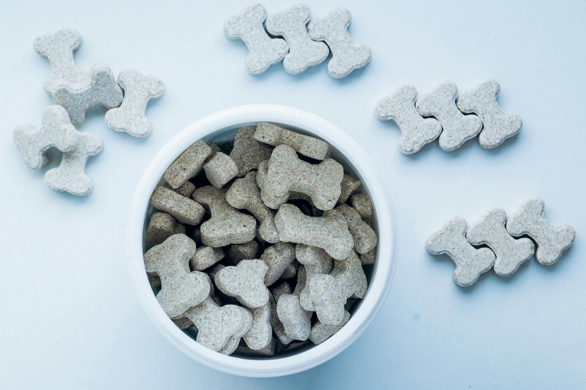 A white bowl filled with bone-shaped dog treats, surrounded by three rows of three treats on a light blue background.