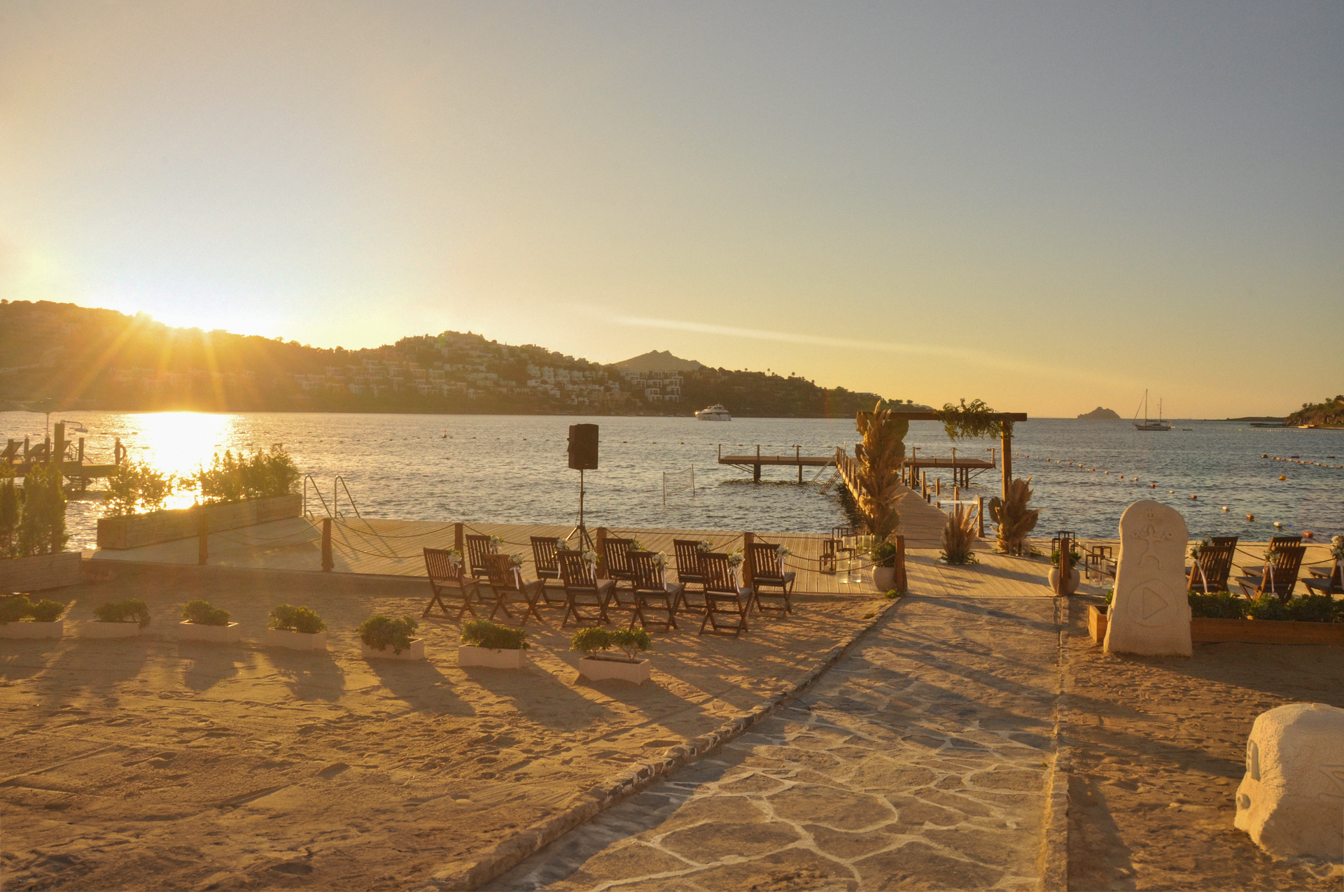 A beach wedding setup with chairs facing a wooden archway by the sea during a golden sunset.