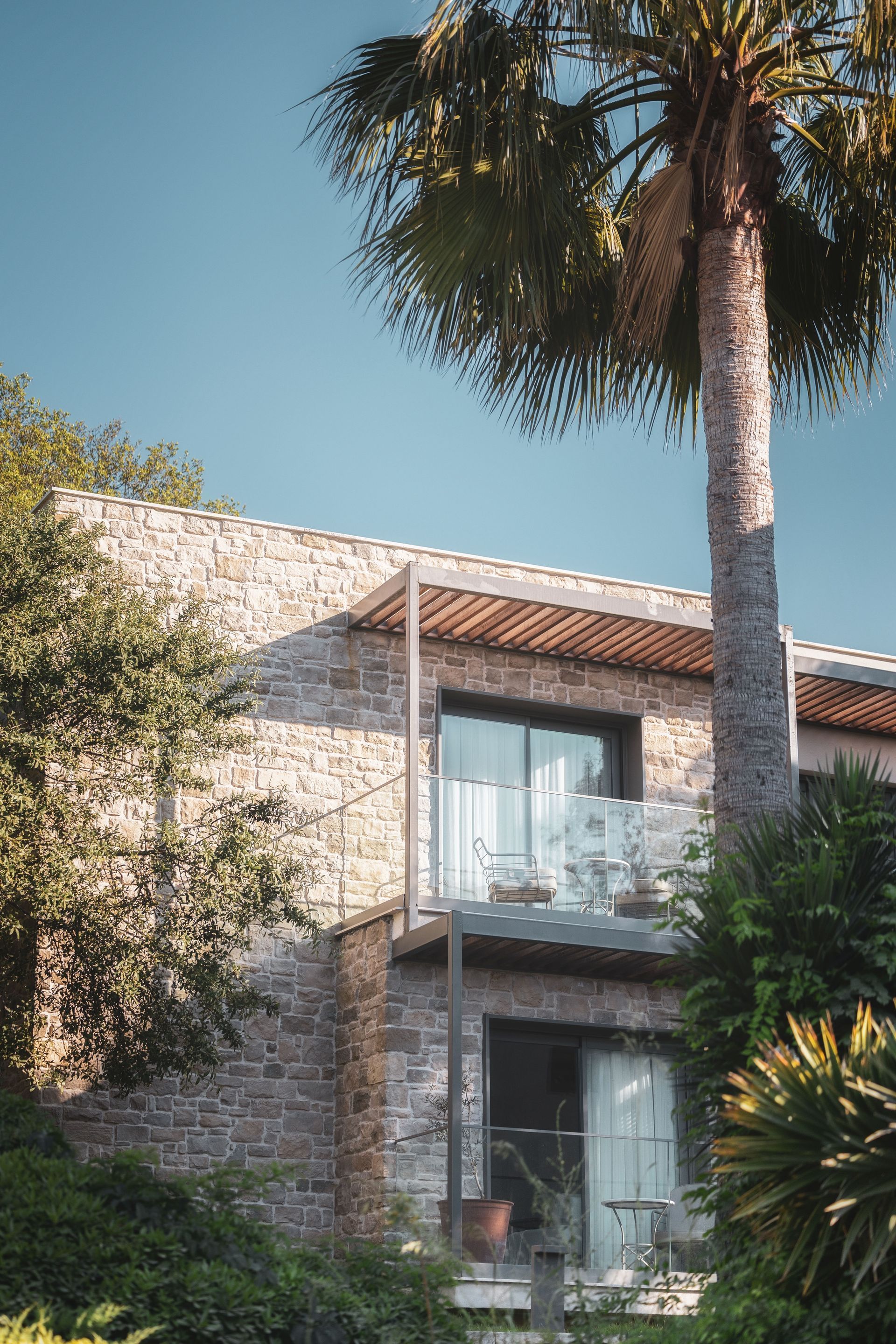 A two-story stone building with glass balcony doors and a pergola, partially shaded by a large palm tree.