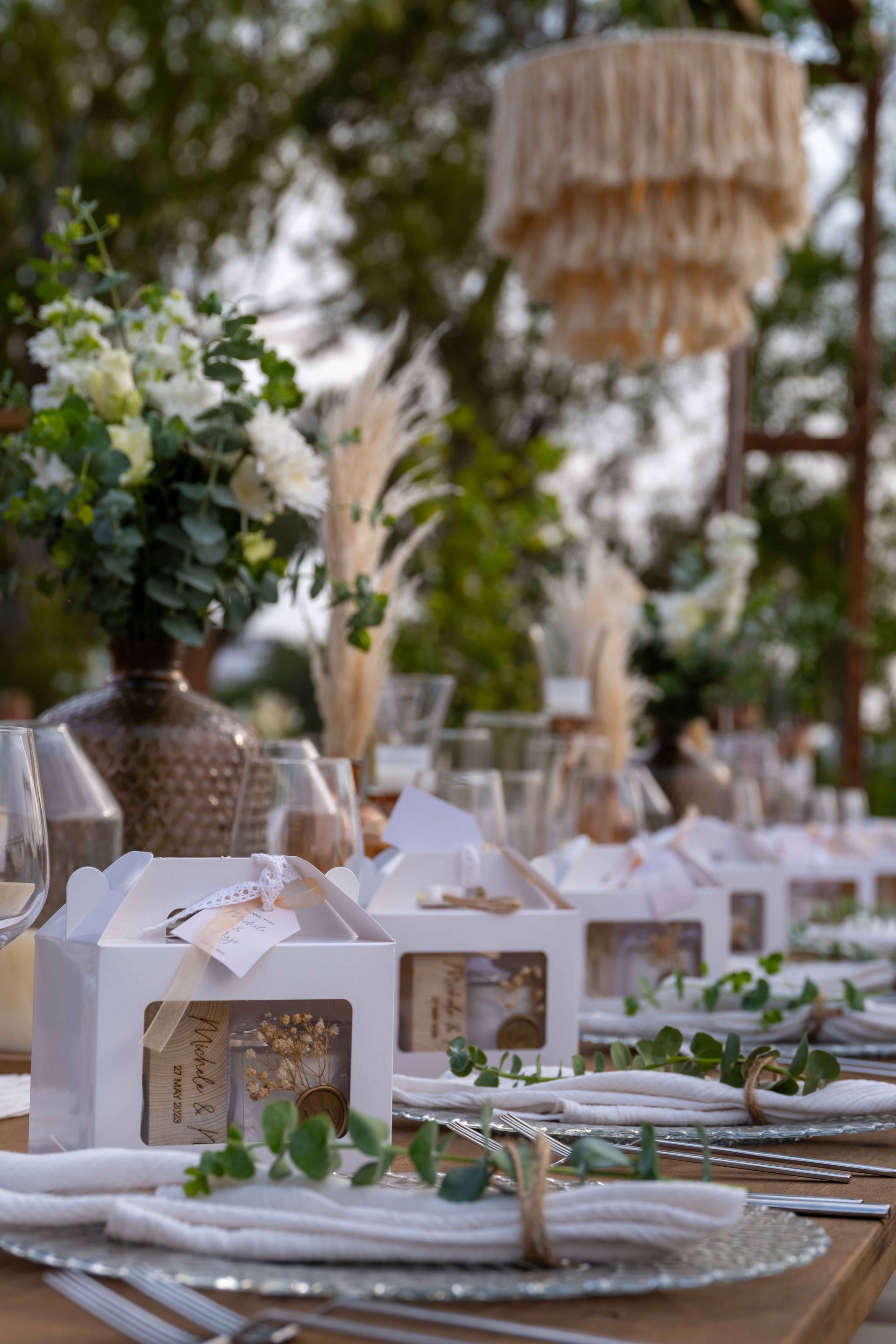 Table setting with white favor boxes, clear plates, napkins with eucalyptus, and boho decor in an outdoor garden setting.