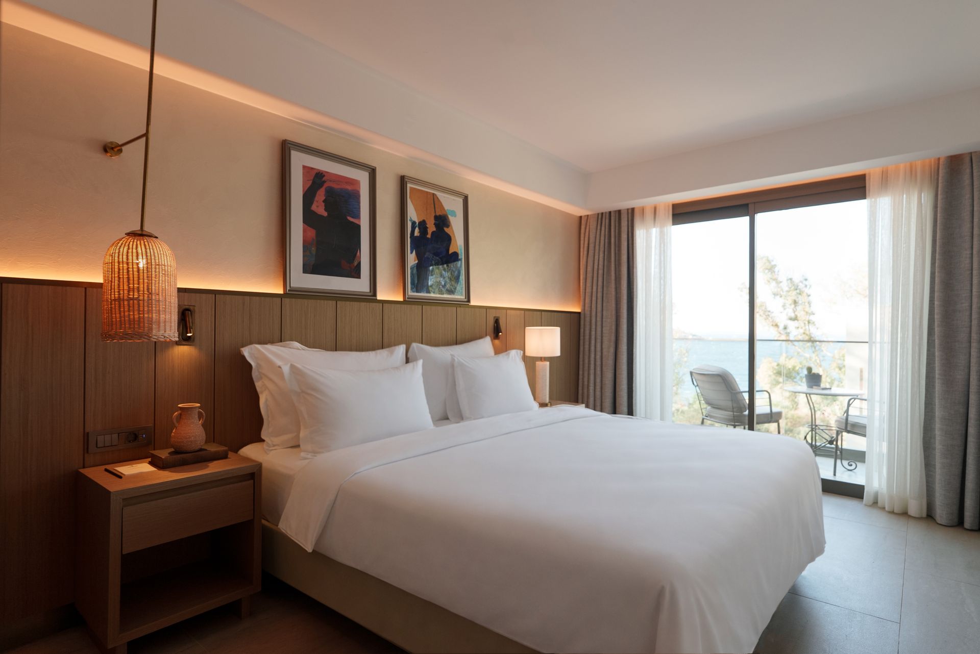 A hotel room with a queen bed, wooden headboard, two pictures, a pendant light, and a balcony view of the sea.