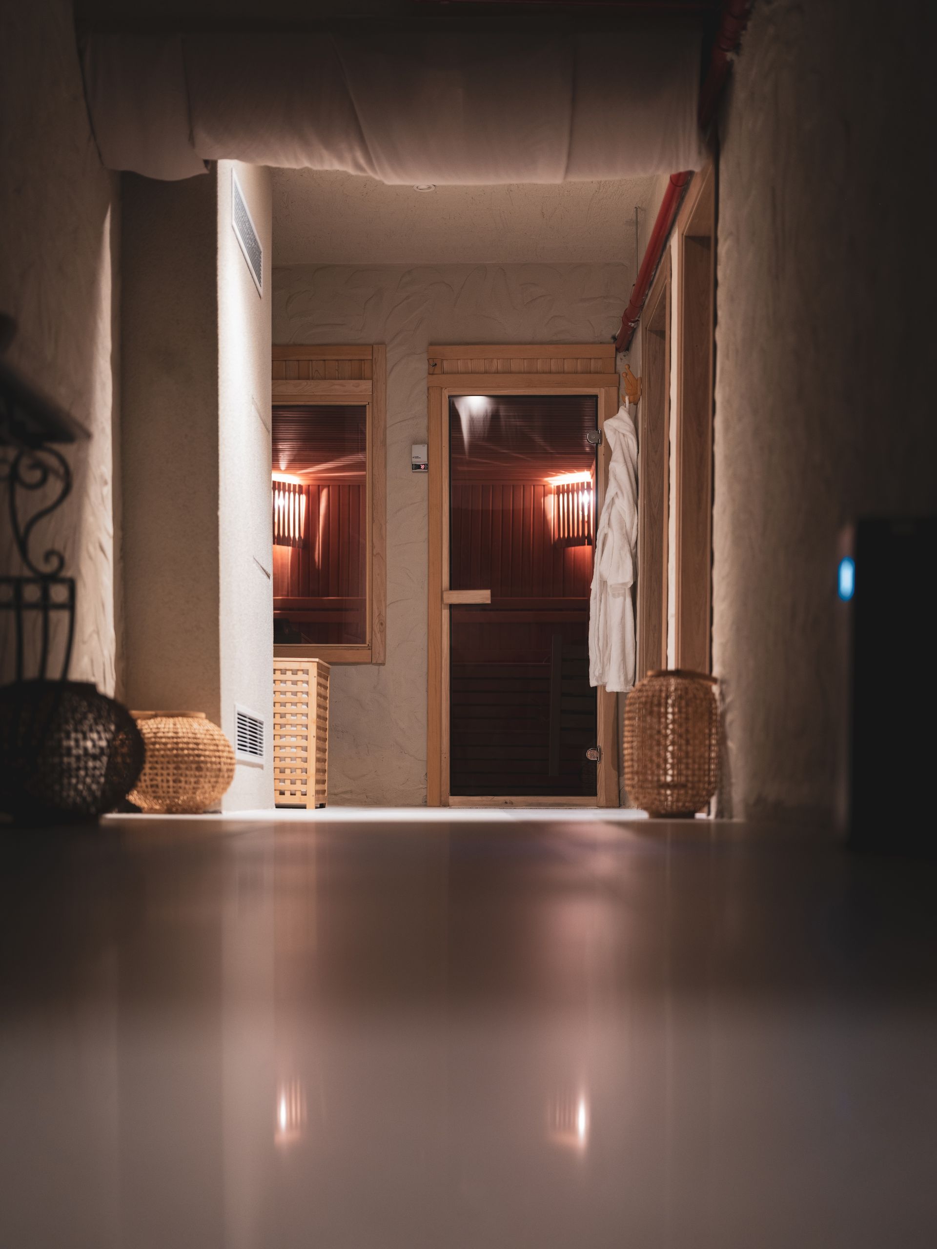A low-angle view of a dimly lit, modern hallway leading to a wooden sauna with warm interior lighting and wicker baskets.