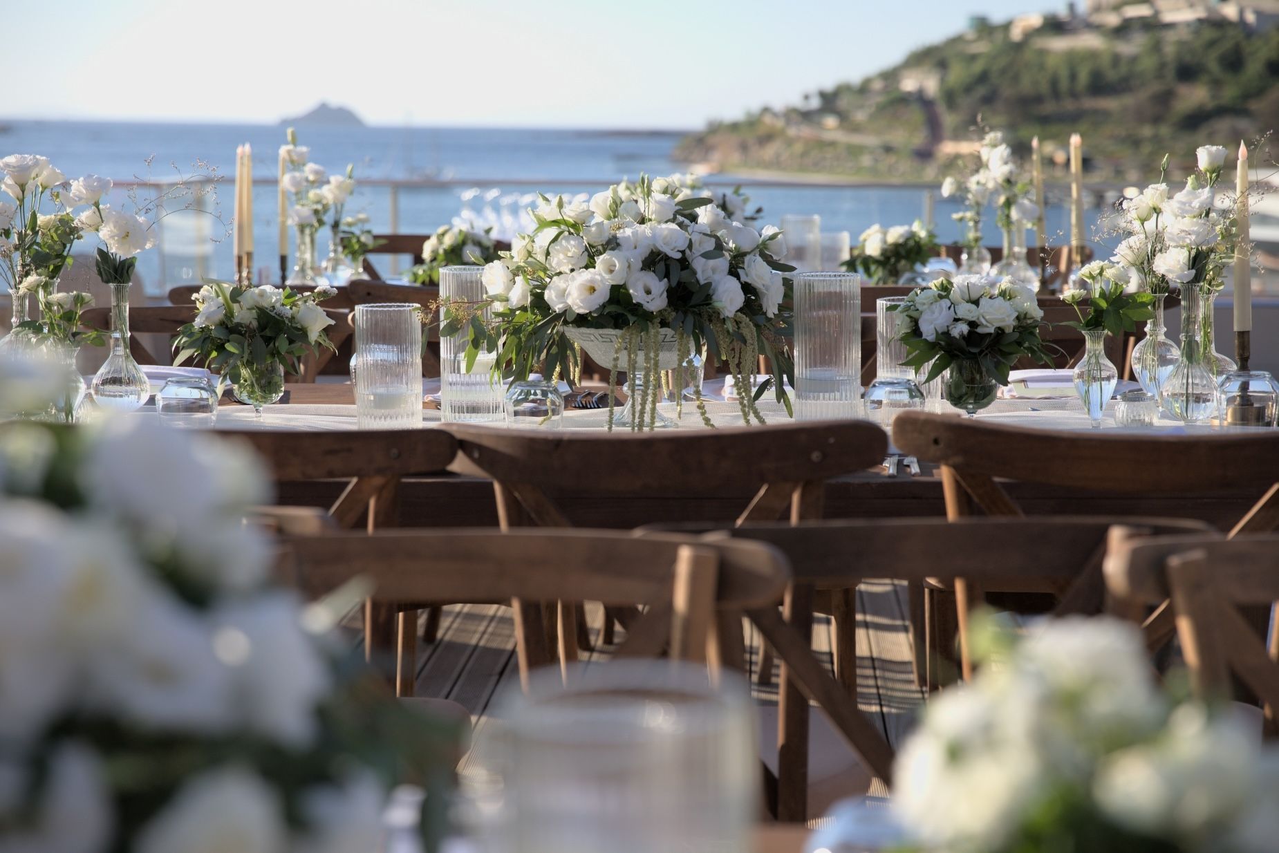 A seaside wedding reception setup with wooden chairs and white flower arrangements on tables overlooking the ocean.