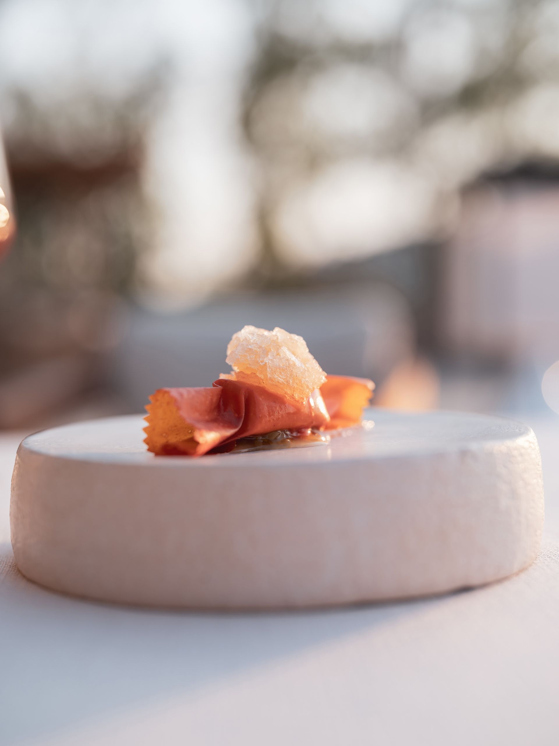 A small, artistic appetizer featuring a folded orange crisp topped with translucent garnish, served on a round white base.