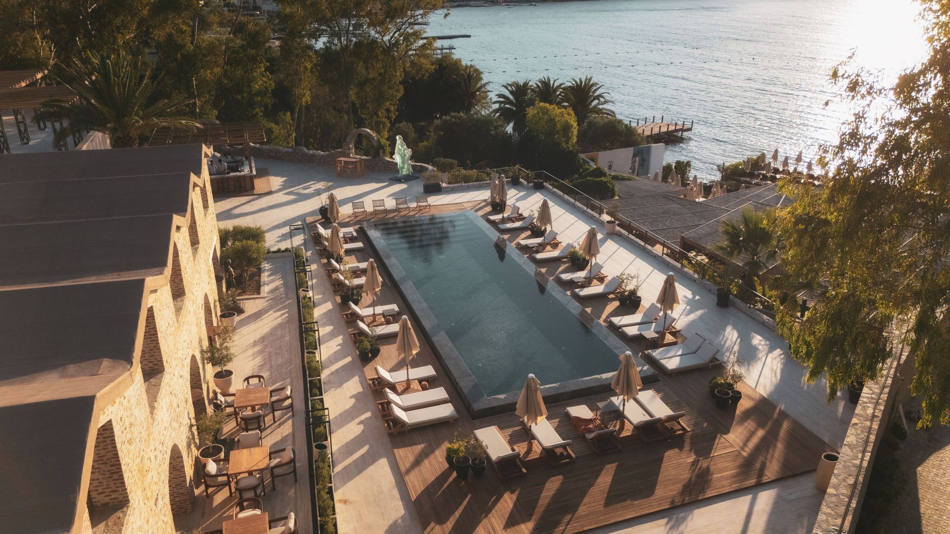 An elevated view of a rectangular infinity pool surrounded by lounge chairs and umbrellas, next to a building by the sea.