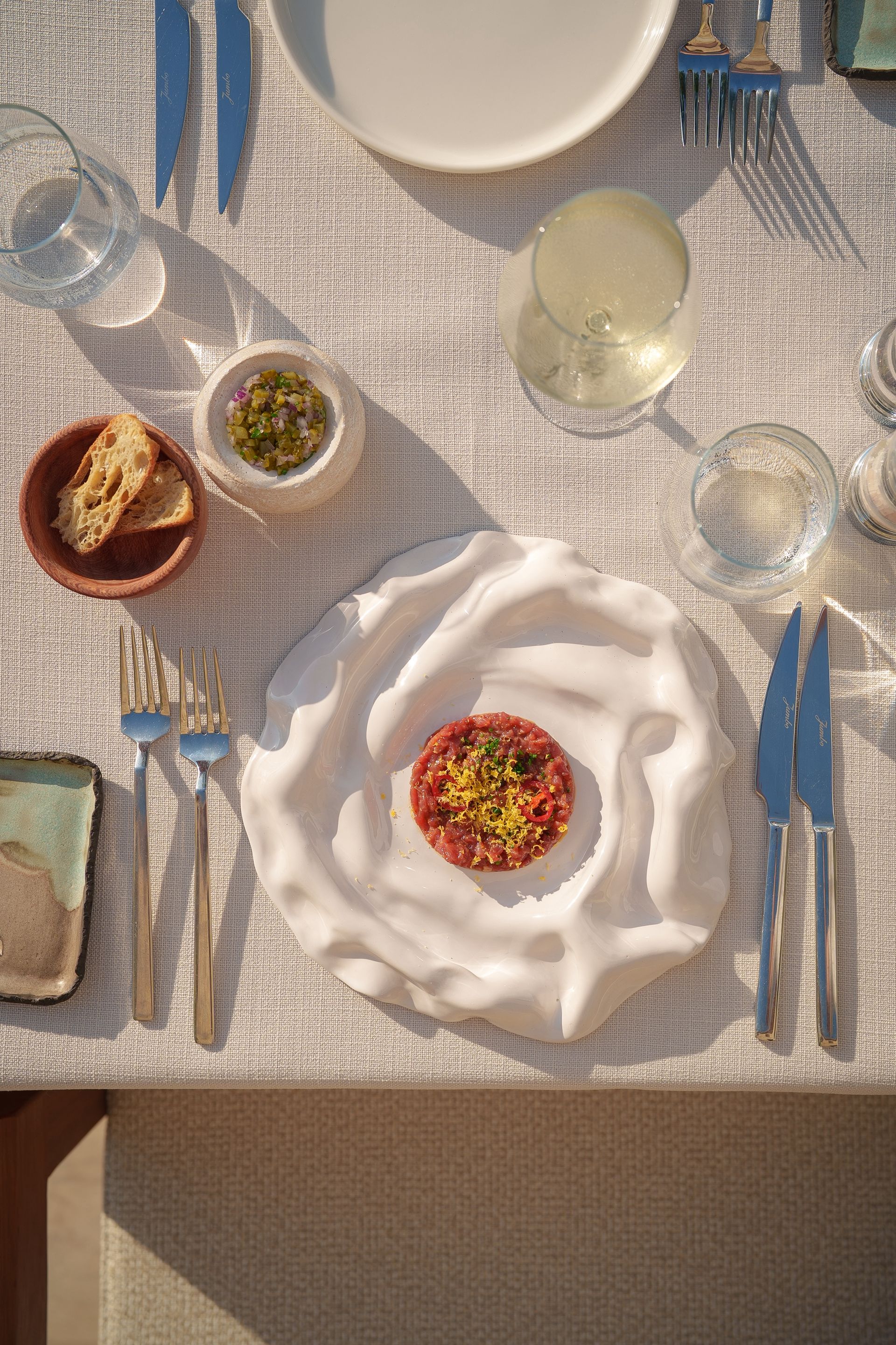 A top-down view of a plate with beef tartare, served on a textured white plate, with cutlery and bread on a white cloth.