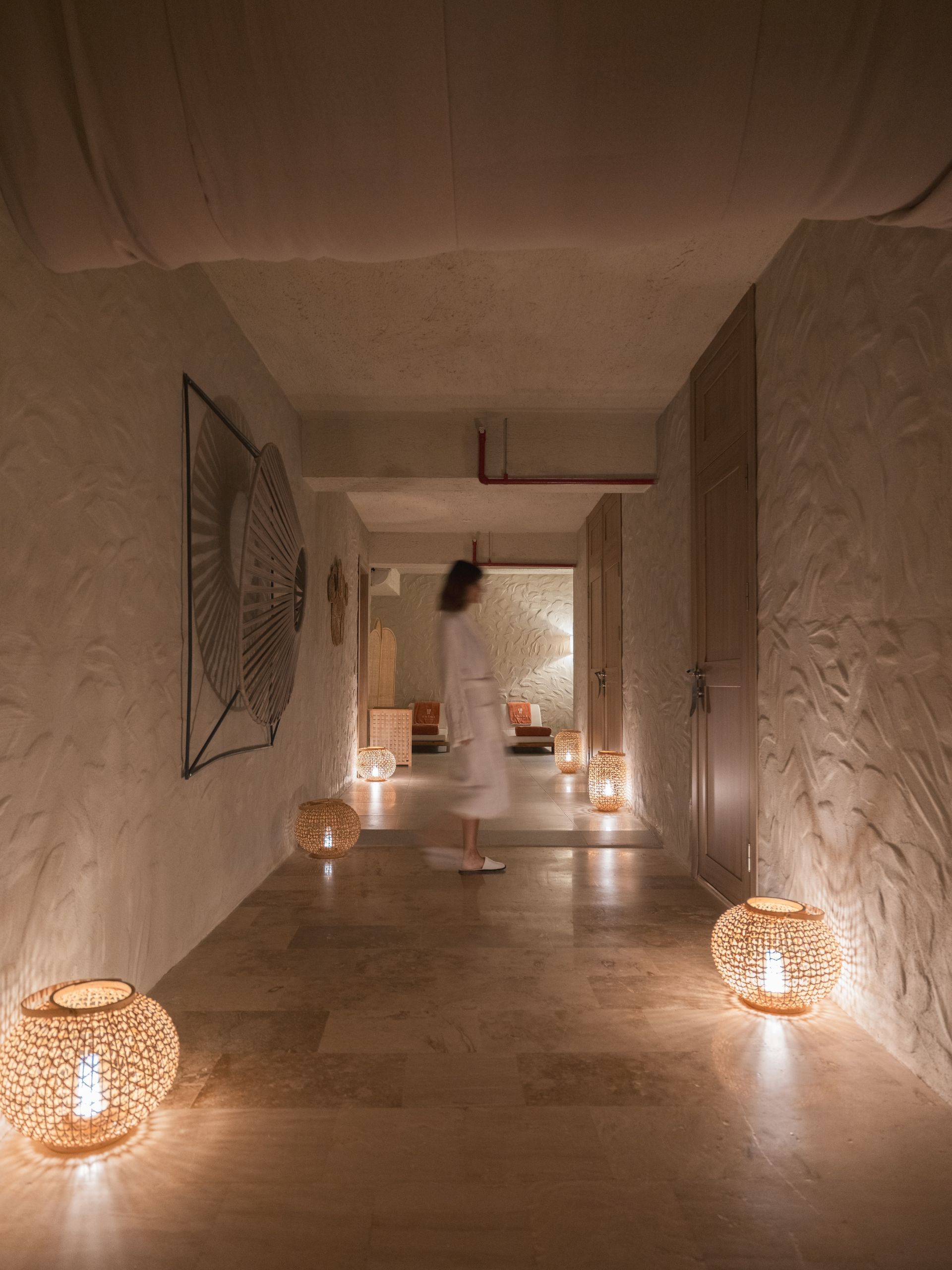 A person in a white robe walks down a dim, stone-walled hallway illuminated by several glowing, intricate floor lamps.