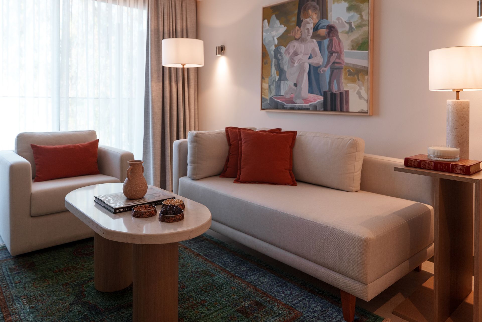 A cream-colored sofa and armchair with rust-colored pillows face a coffee table, with a painting above the couch.