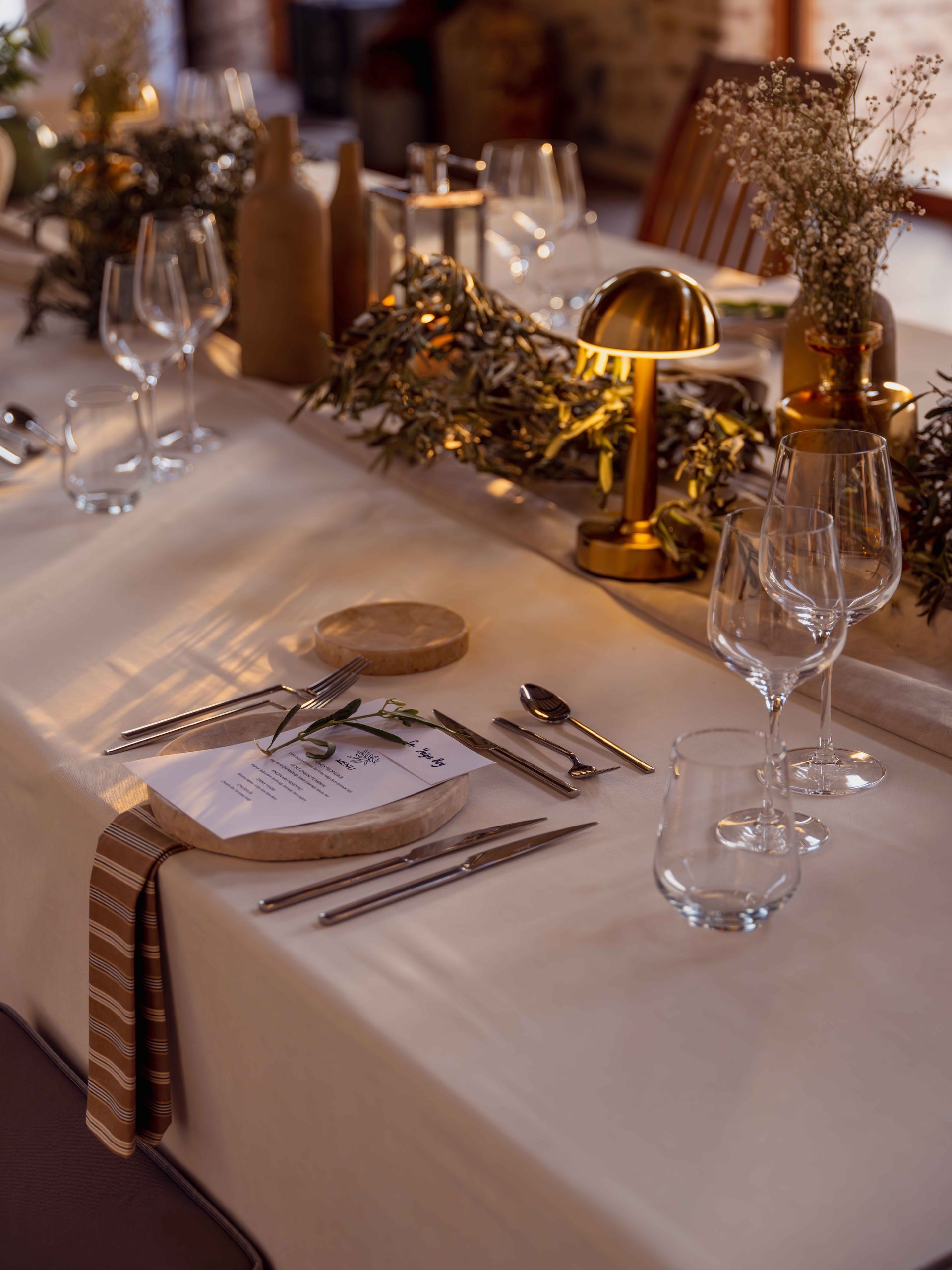 A warm, elegant table setting featuring beige linens, delicate glassware, gold lamps, and greenery for a formal event.