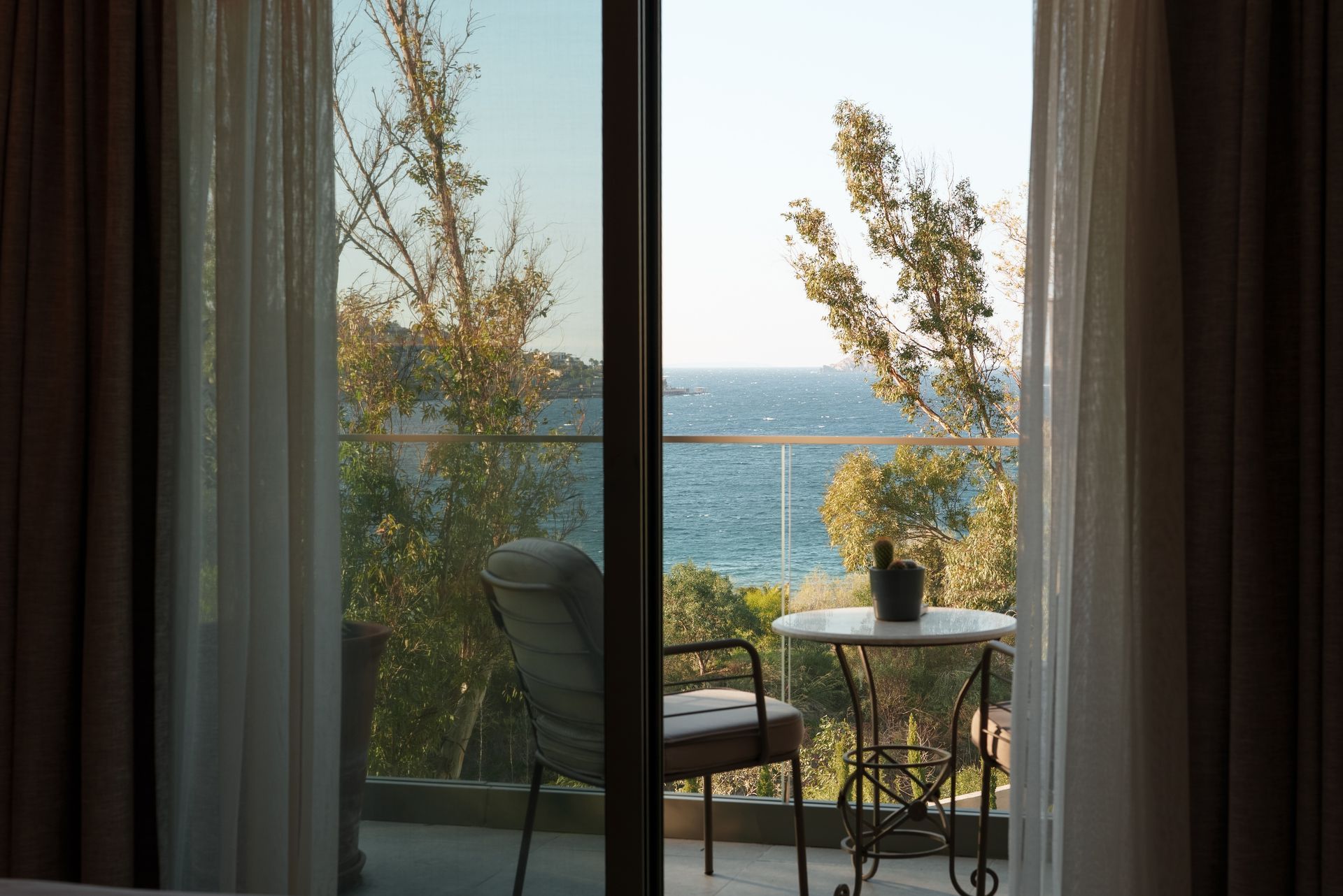 A view through an open balcony door showing a chair, a small table, trees, and the ocean under a clear sky.