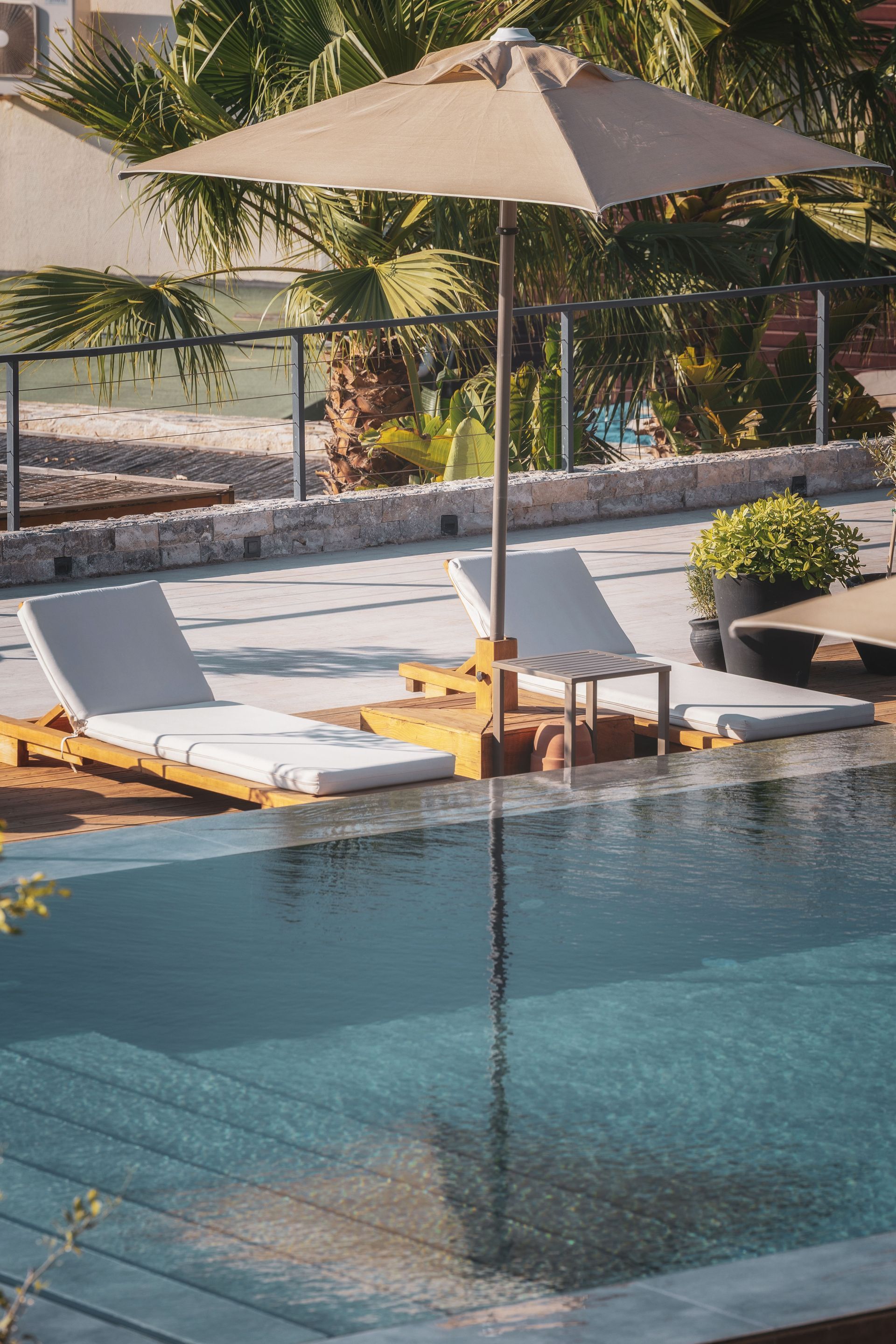 Two lounge chairs and a tan umbrella by a sparkling outdoor swimming pool on a sunny day.