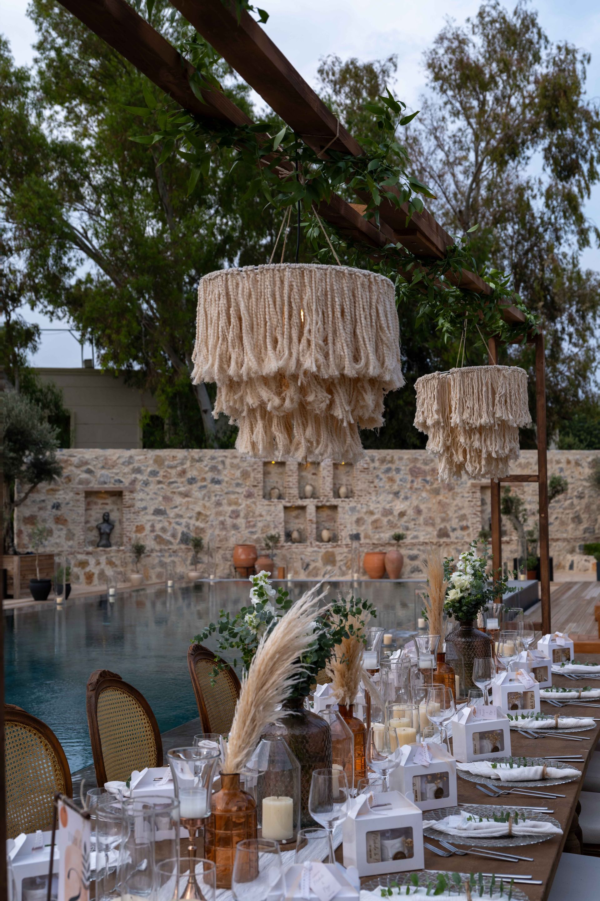 Outdoor dinner table set for an event beside a pool, featuring hanging macramé chandeliers and pampas grass centerpieces.