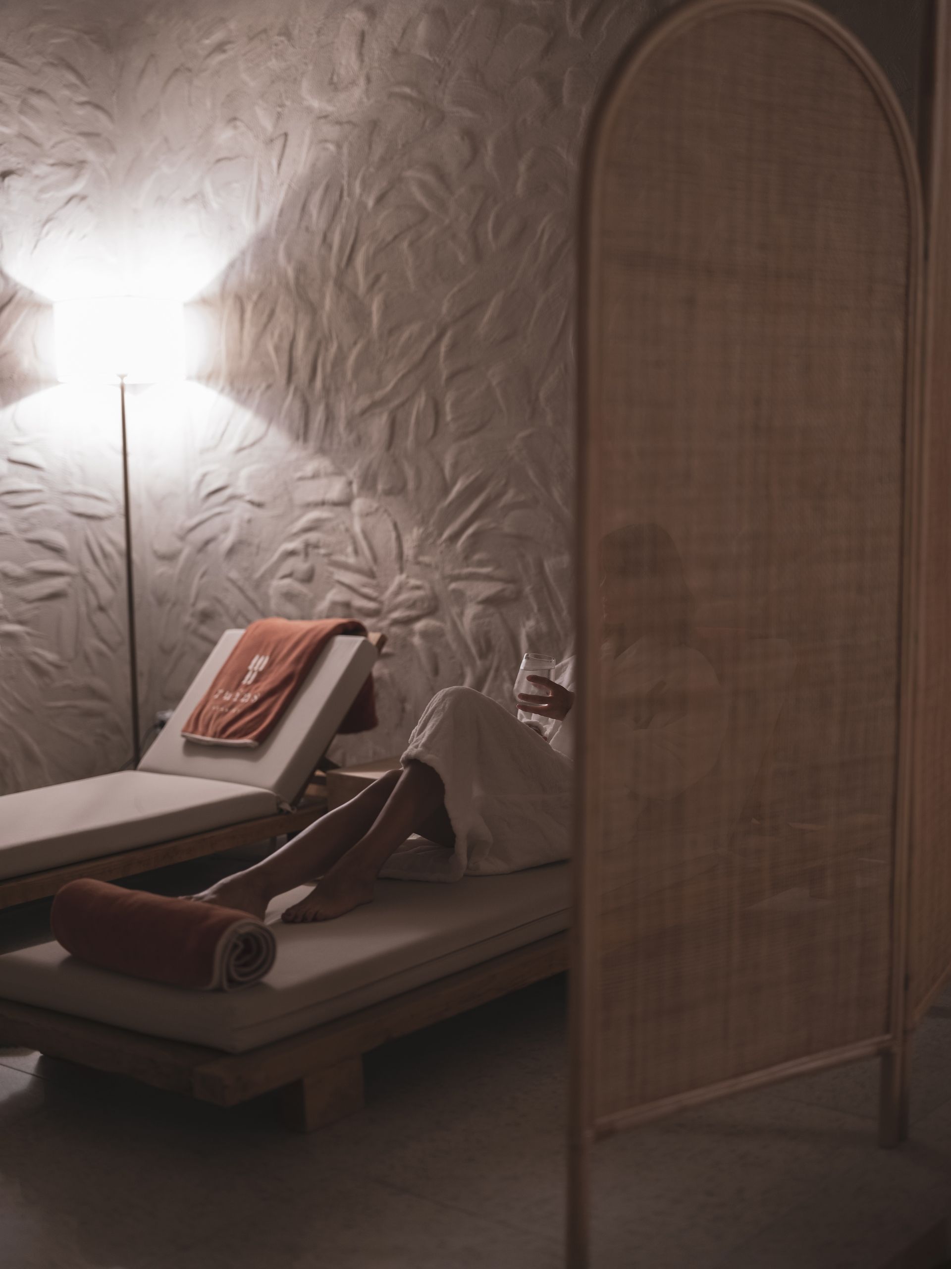 A person relaxes on a spa lounge chair behind a curved rattan screen in a dimly lit room with textured walls.