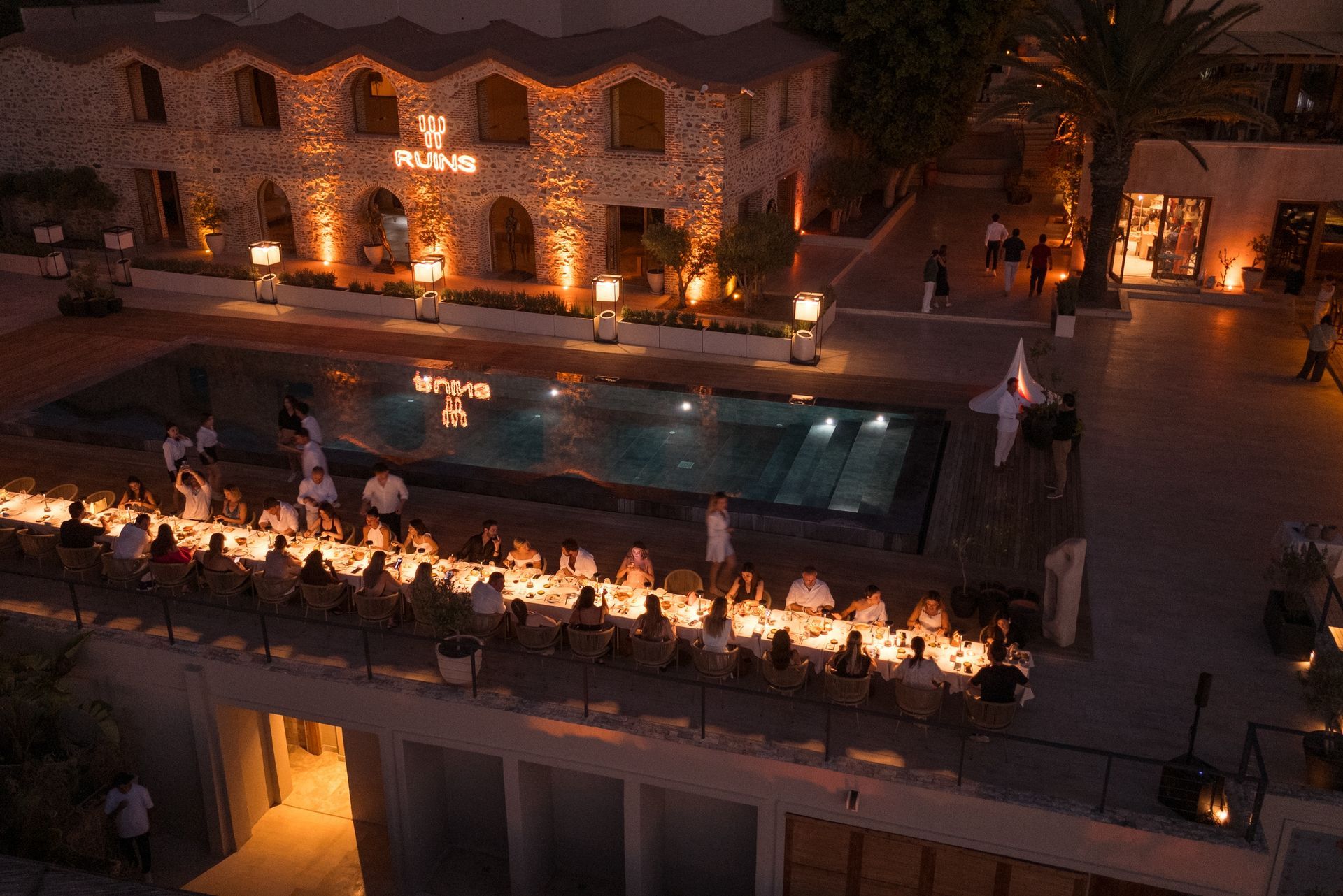An overhead view of a nighttime dinner party with a long table, glowing lights, and a pool set against a stone building.