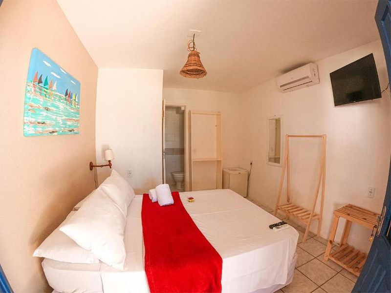 Quarto com cama, TV, obras de arte e banheiro privativo. Paredes brancas, peseira vermelha e móveis de madeira clara.