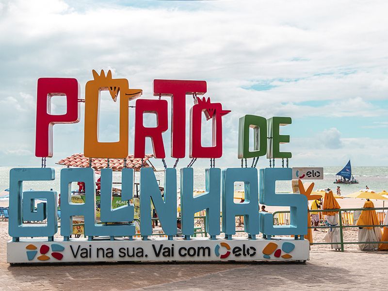 Placa colorida com a inscrição "Porto de Galinhas" na praia. Água azul, céu nublado, guarda-sóis amarelos e pessoas ao fundo.