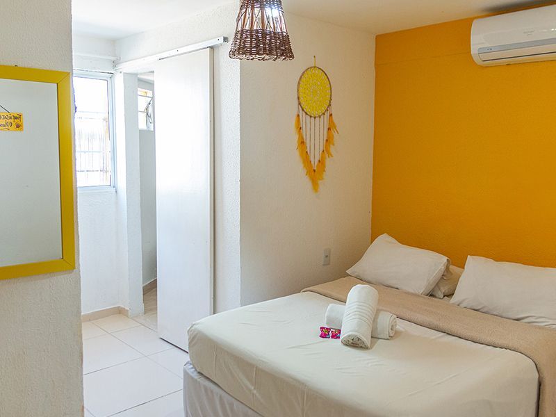 Quarto com cama, parede com detalhes em amarelo, espelho, filtro dos sonhos e ar condicionado.