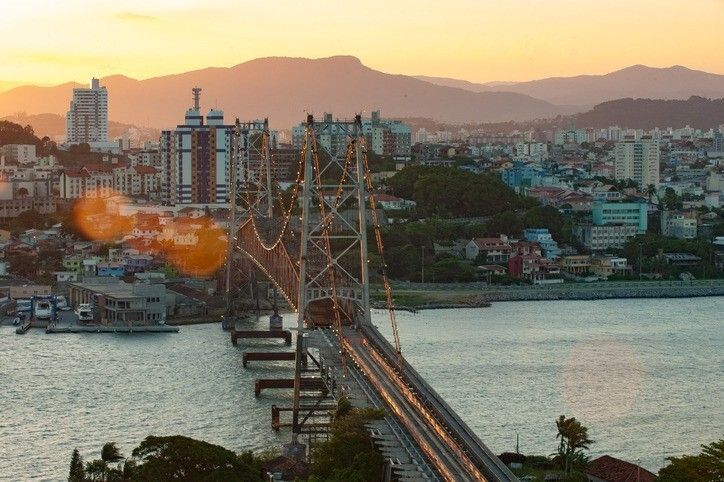 Ponte suspensa sobre a água ao pôr do sol, com a cidade e as montanhas ao fundo. Tons de laranja e dourado.