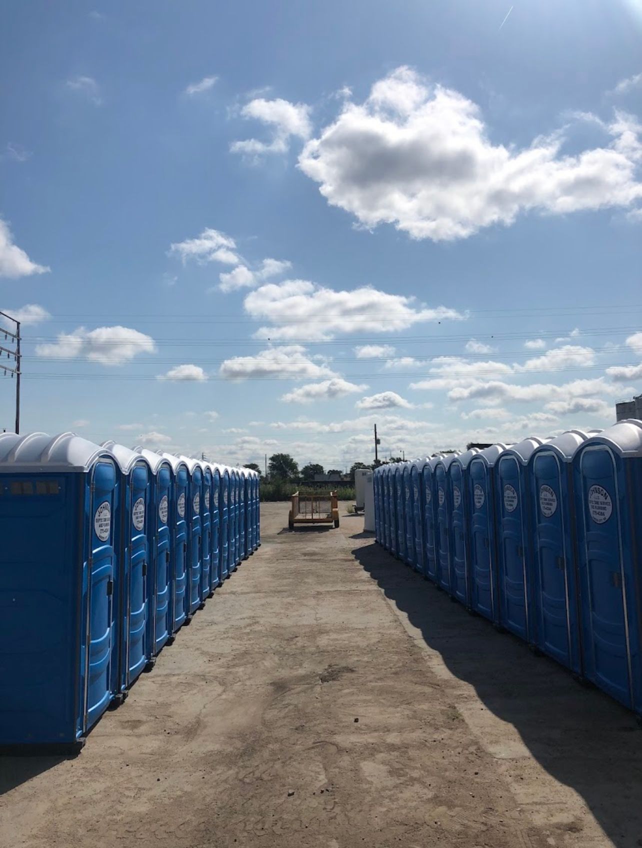 Rows of blue portable toilets line a concrete lot under a partly cloudy sky. A small vehicle is parked between them.