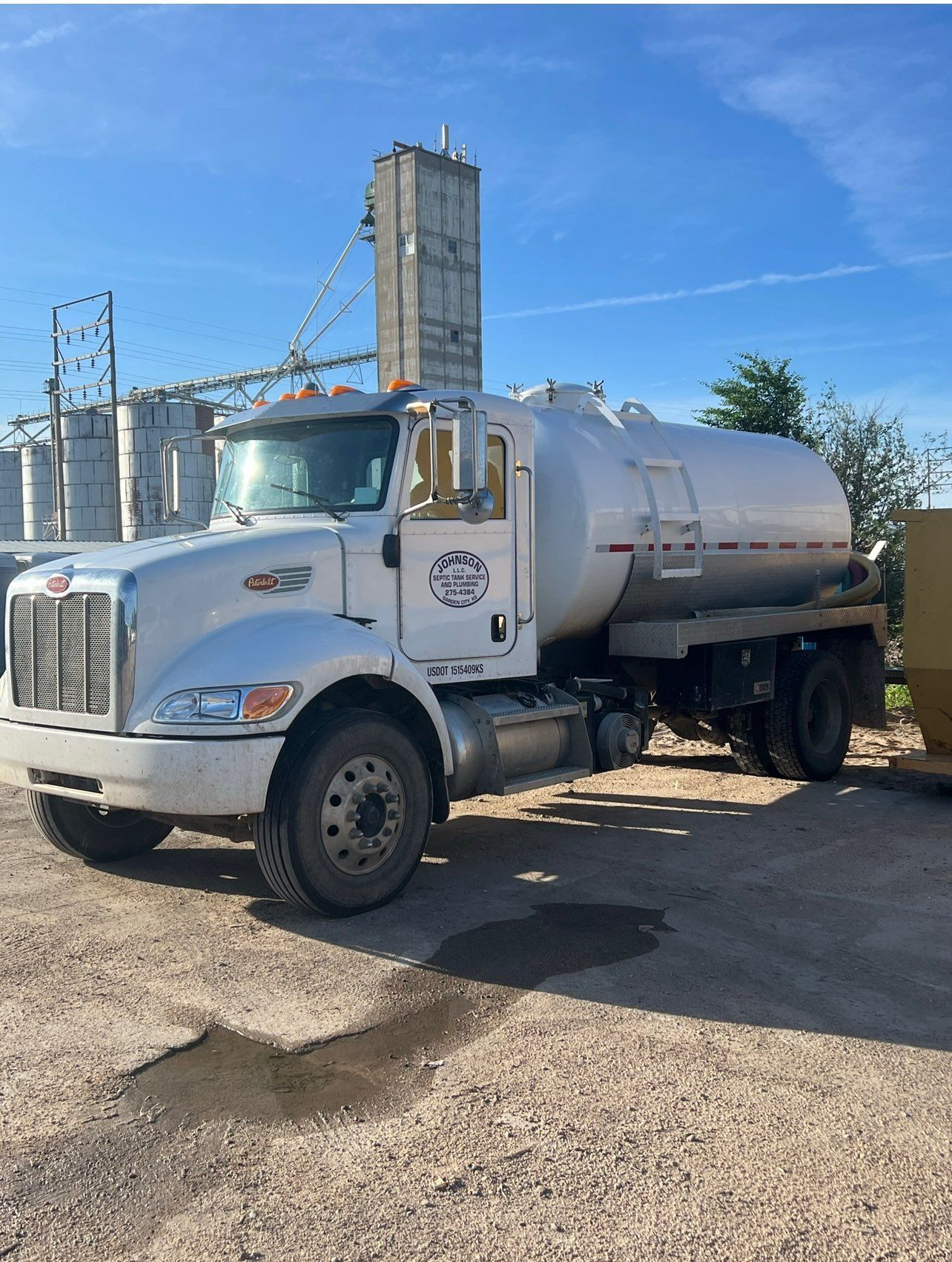 White tanker truck parked outdoors near silos on a sunny day.