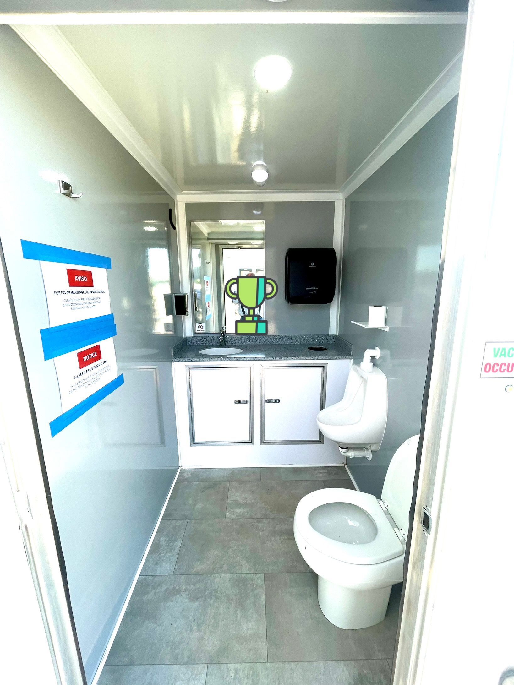 Interior of a portable restroom with a toilet, sink, and mirror. Gray walls and floor.