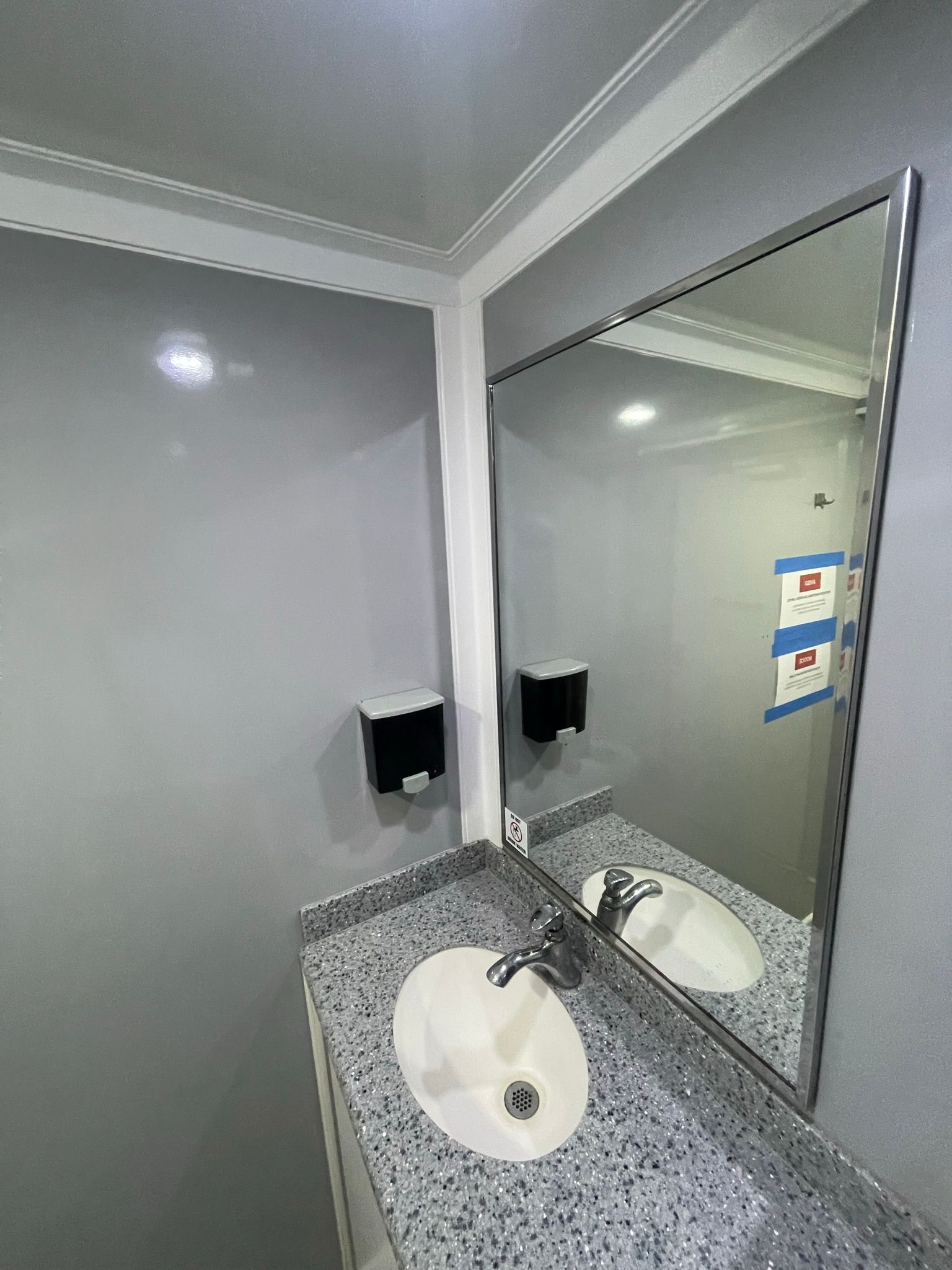 A public bathroom with a sink and mirror, grey walls, and a black soap dispenser.