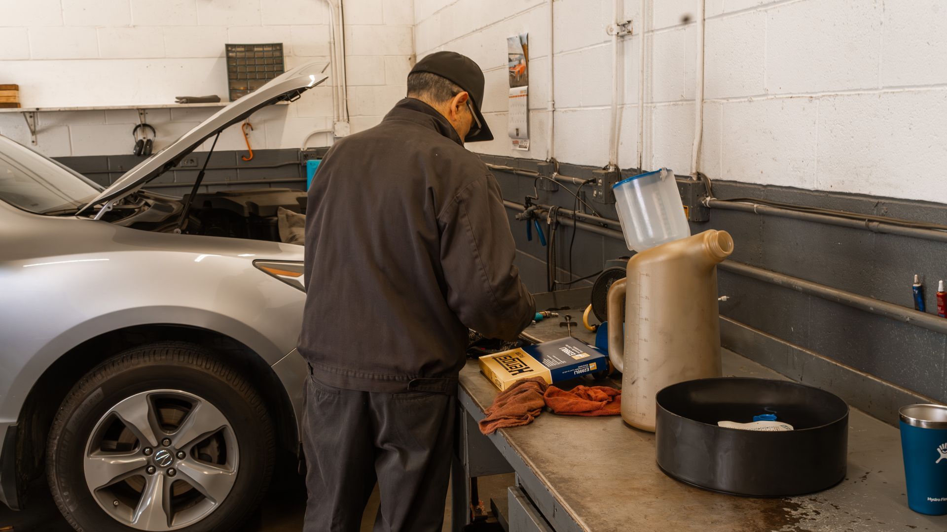 Mechanic working on a car in a garage. Car is silver, workbench has tools and containers.