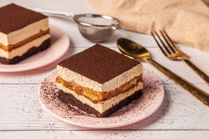 Un pezzo di tiramisù è su un piatto rosa accanto a una forchetta e un cucchiaio.