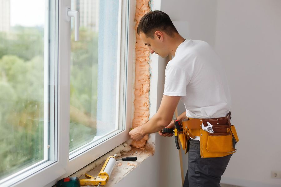 Persona che installa materiale isolante accanto a una finestra in una stanza luminosa, utilizzando una cintura portautensili e guanti da lavoro.