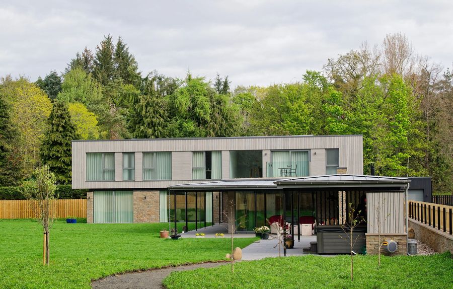 Casa moderna a due piani con ampie vetrate, affacciata su un prato verde e un patio circondato da alberi.
