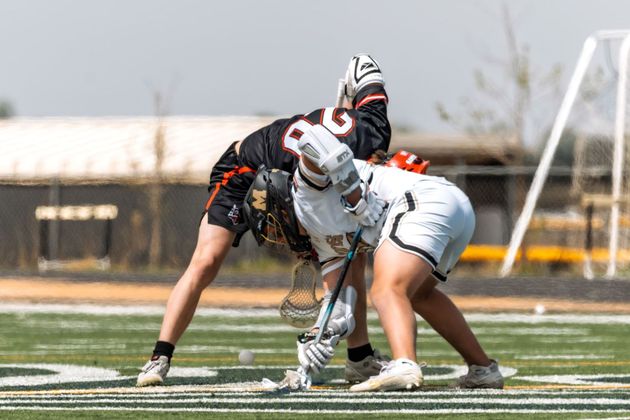 Two lacrosse players in white and black uniforms face off on a field, crouched over the ball.