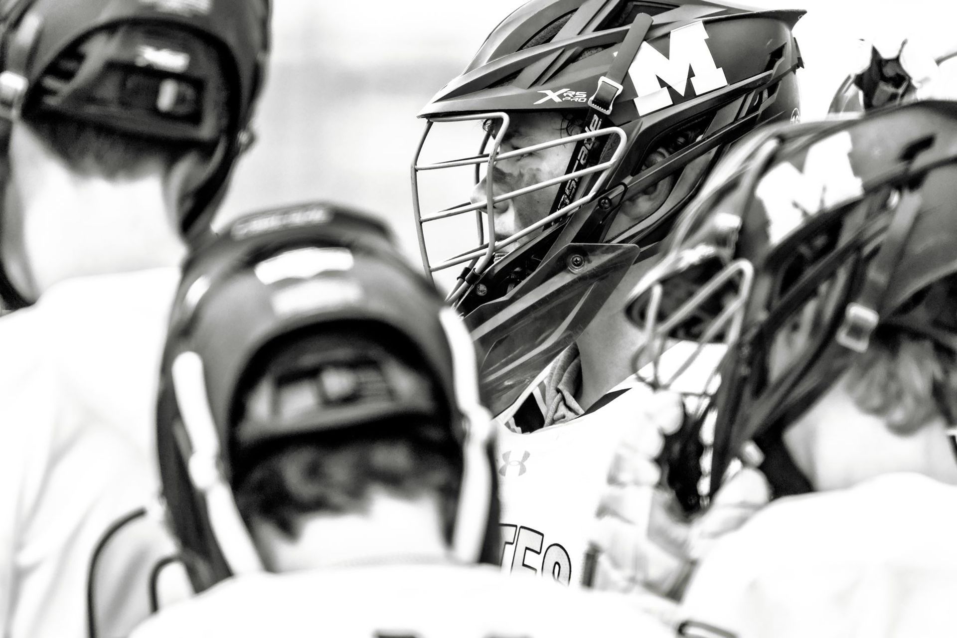 Lacrosse players in helmets and uniforms huddle on the field, seen in black and white.