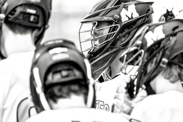Lacrosse players in helmets and uniforms huddle on the field, seen in black and white.