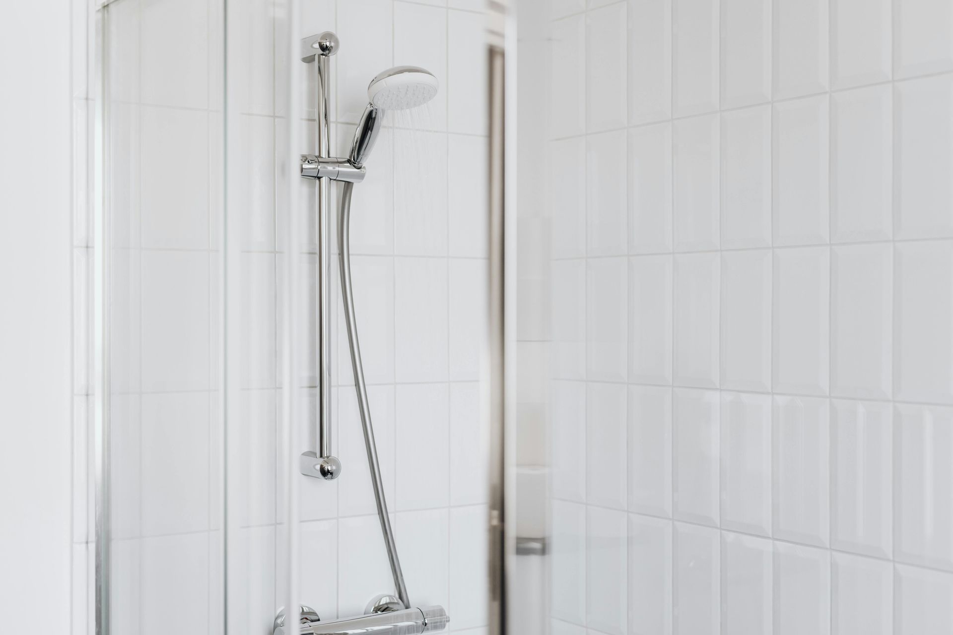 A shower head and silver hose attached to a vertical sliding bar, mounted against a wall of white rectangular tiles.