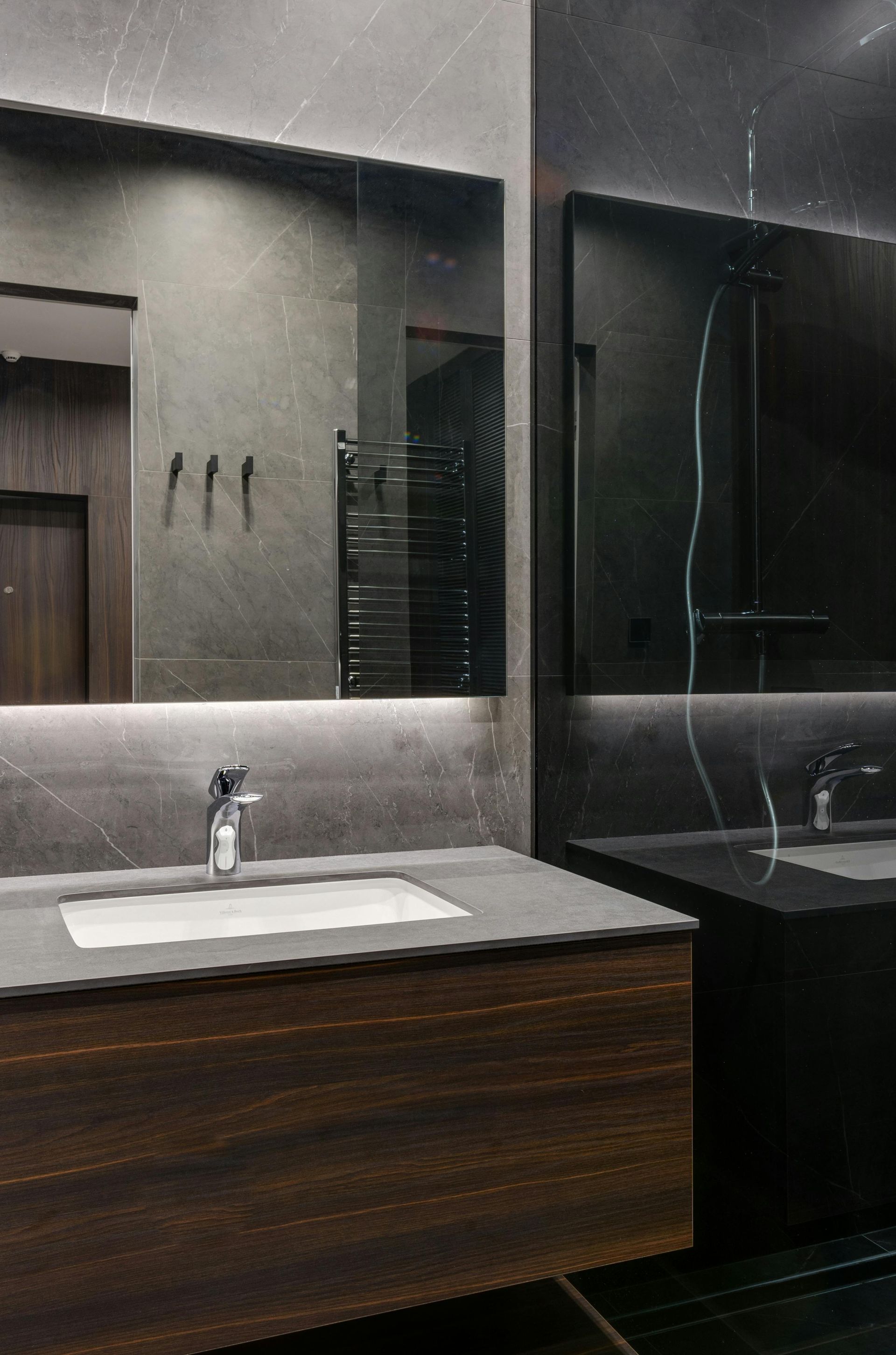 Modern bathroom with a wooden floating vanity, white sink, gray stone wall, large rectangular mirror, and dark fixtures.
