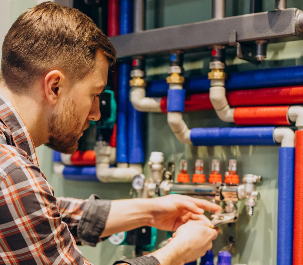 A professional in a plaid shirt uses a wrench to adjust plumbing valves on a wall-mounted heating system with colored pipes.