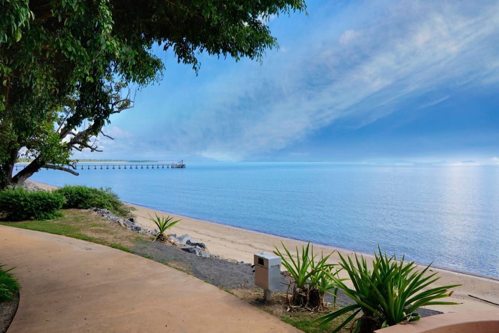 Sunny Beach Walkway Beside Calm Blue Water, With Trees, Plants, and a Distant Pier — Cairns Floors and More In Cardwell, QLD
