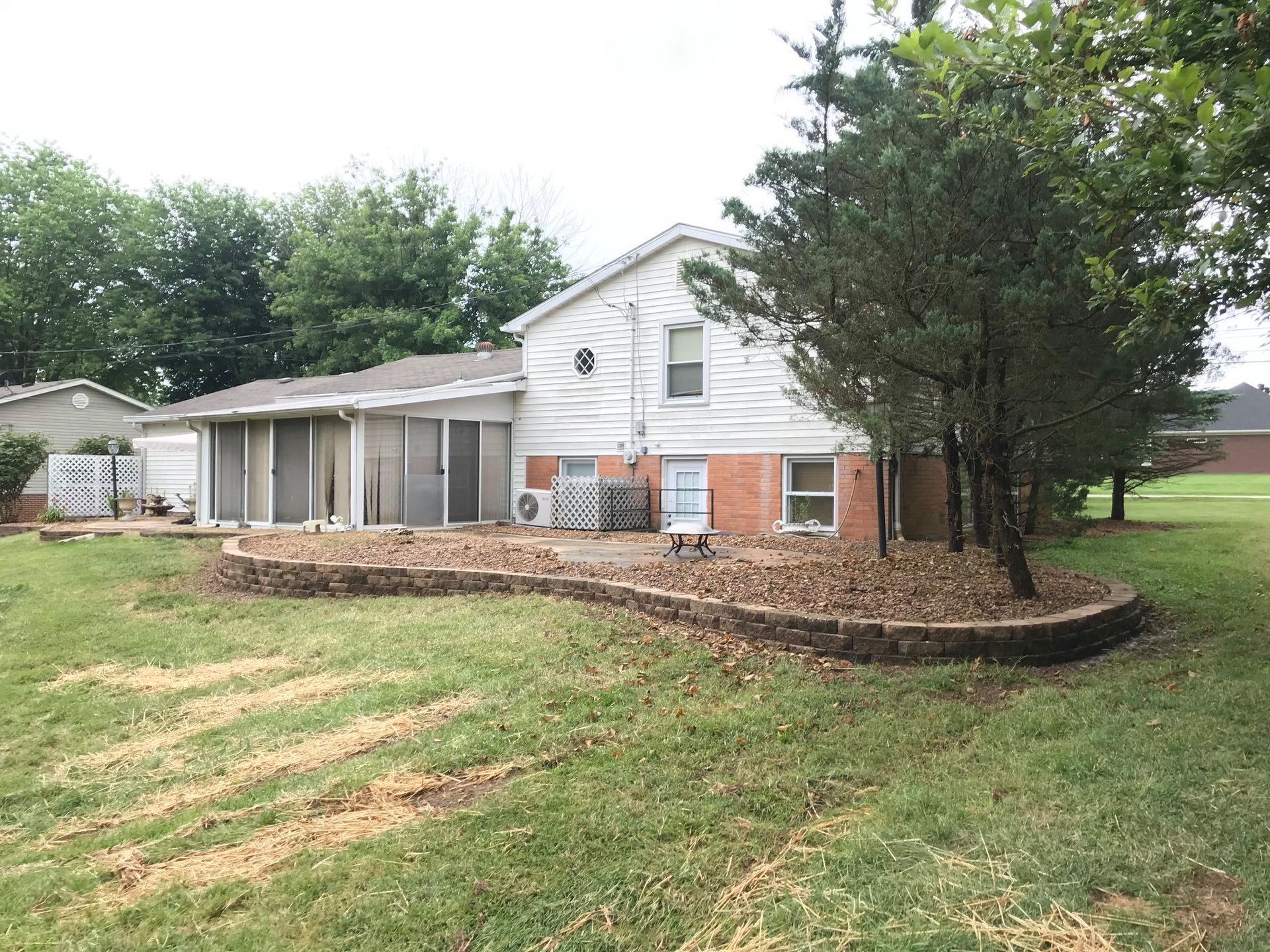 White house with covered porch and brick foundation beside a large tree and grassy yard