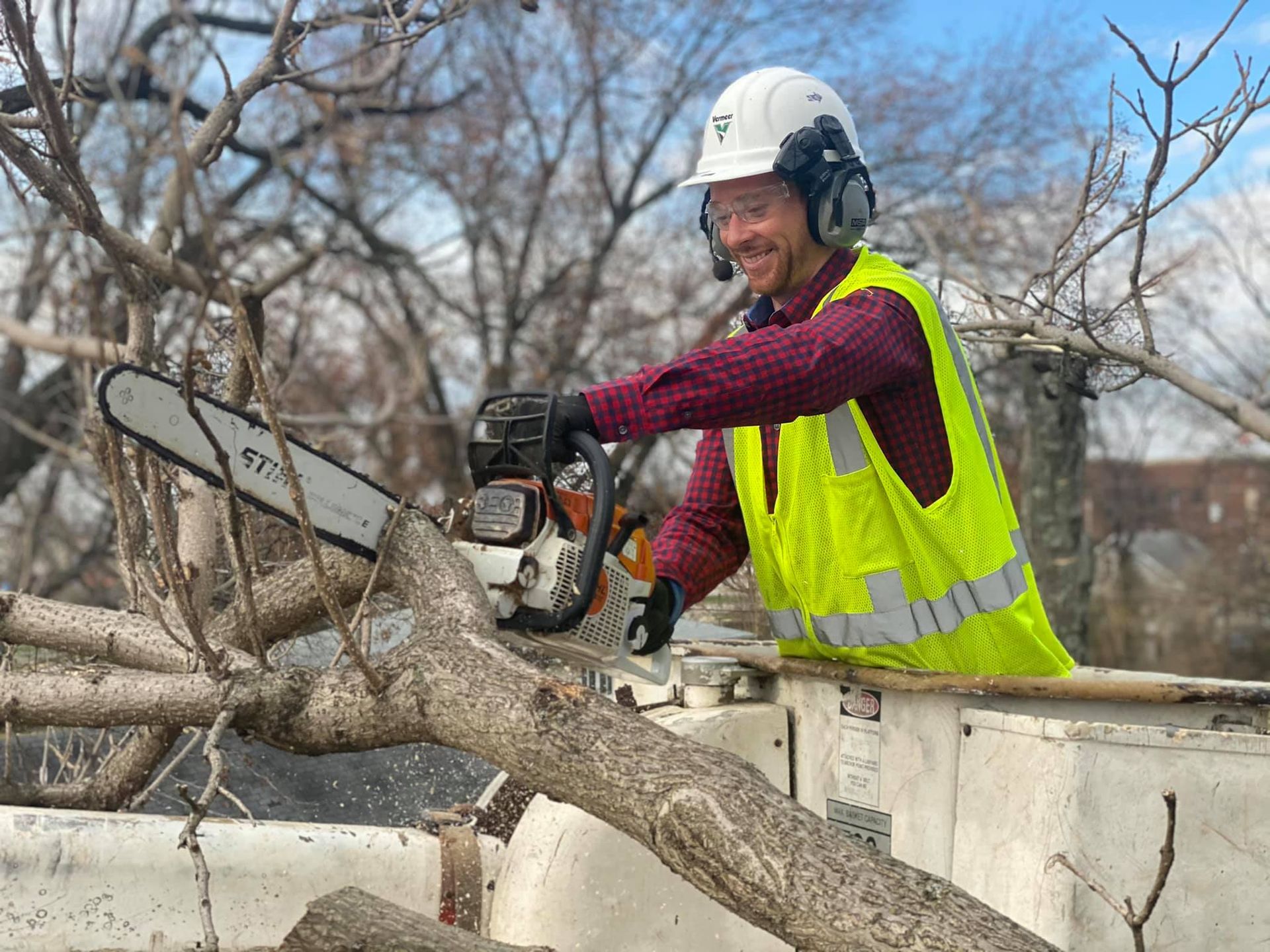 Worker using a chainsaw to cut a tree branch from a bucket truck, wearing a hard hat and safety vest