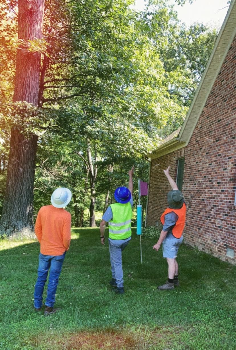 Three children in hard hats and safety vests walk along a grassy path beside buildings and trees.